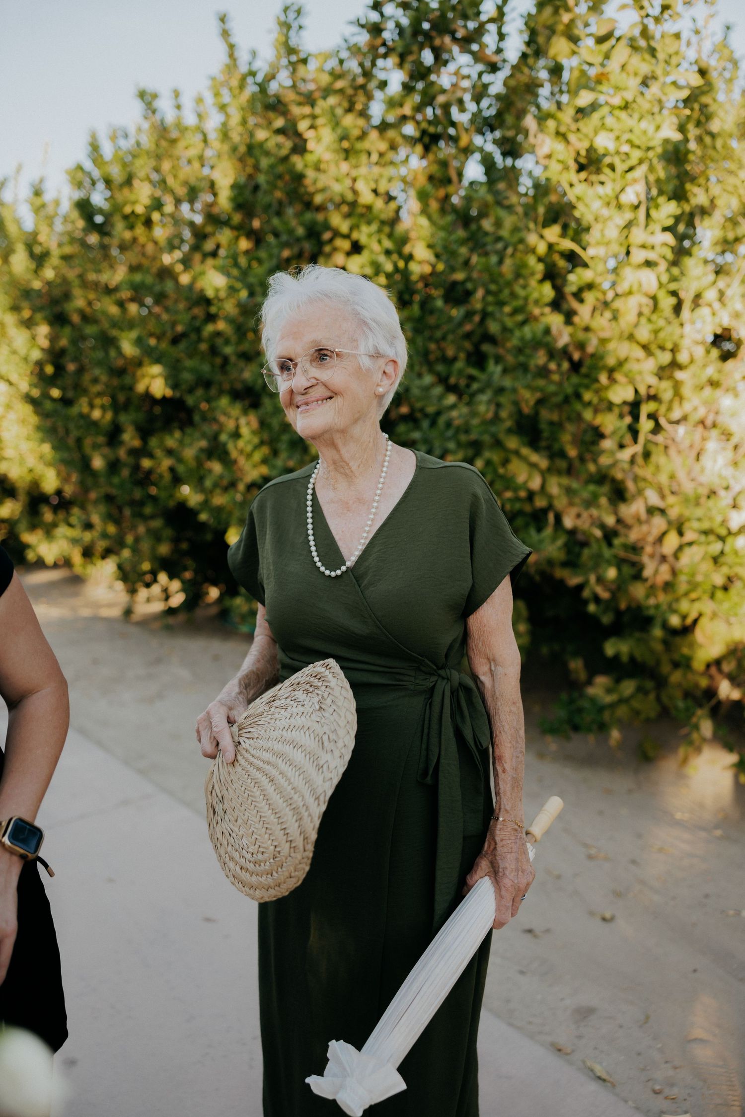 Woman in black dress with white gloves and woven fan smiles while standing near citrus trees in garden setting.