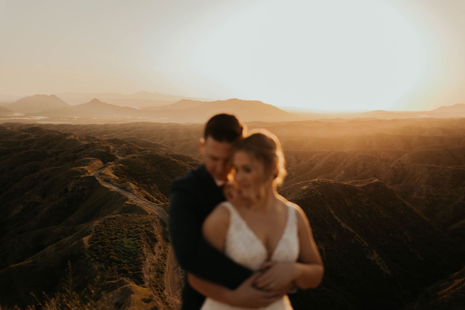 Elegant couple in wedding attire embraces against scenic mountain backdrop during golden sunset.