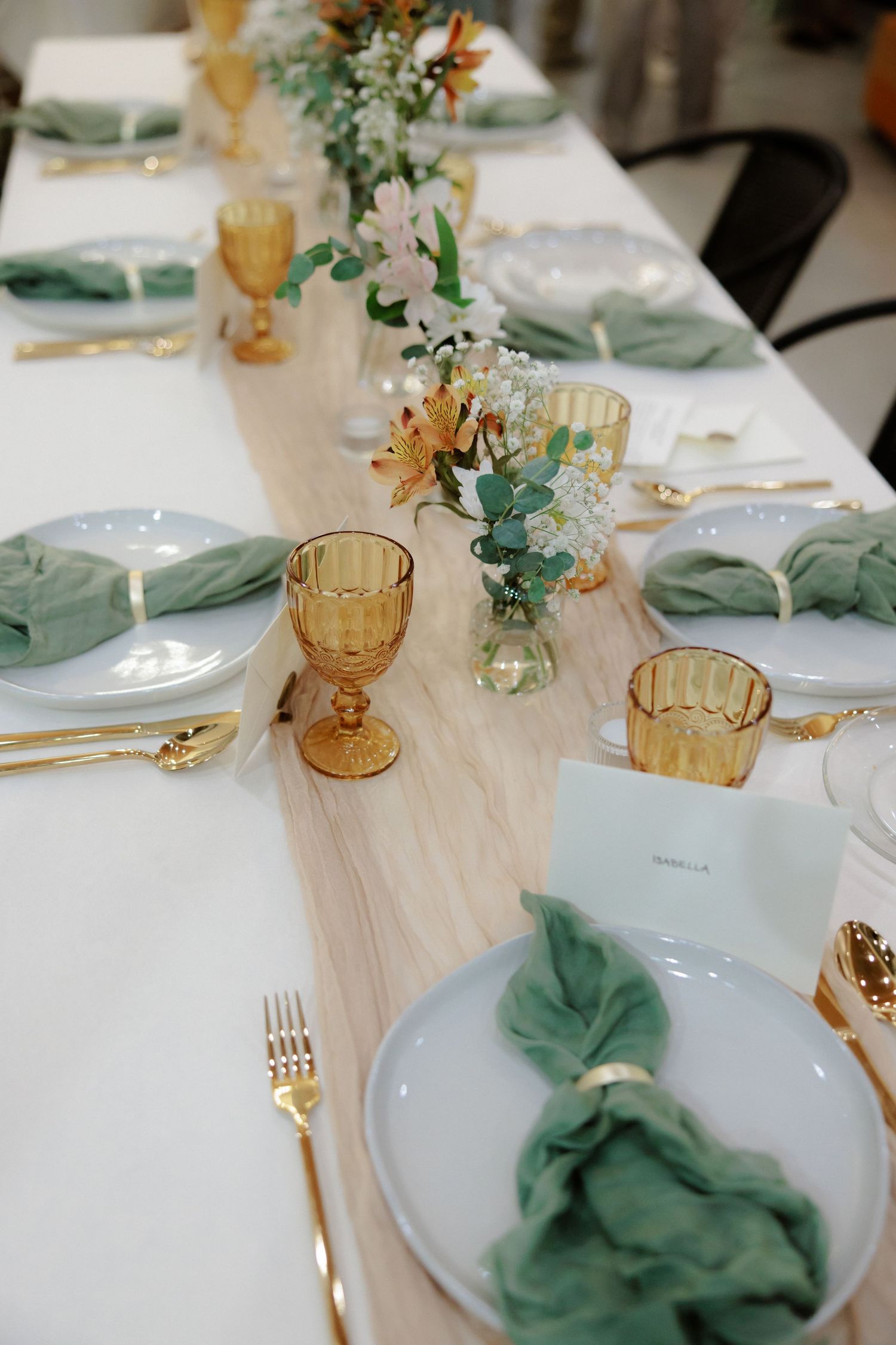 Elegant table setting with sage green napkins, amber glassware, and white plates on a light linen tablecloth.