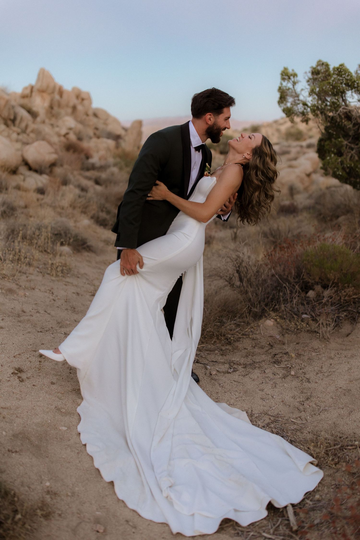 A romantic couple in wedding attire share an intimate moment at sunset in a desert landscape with rocky outcroppings.