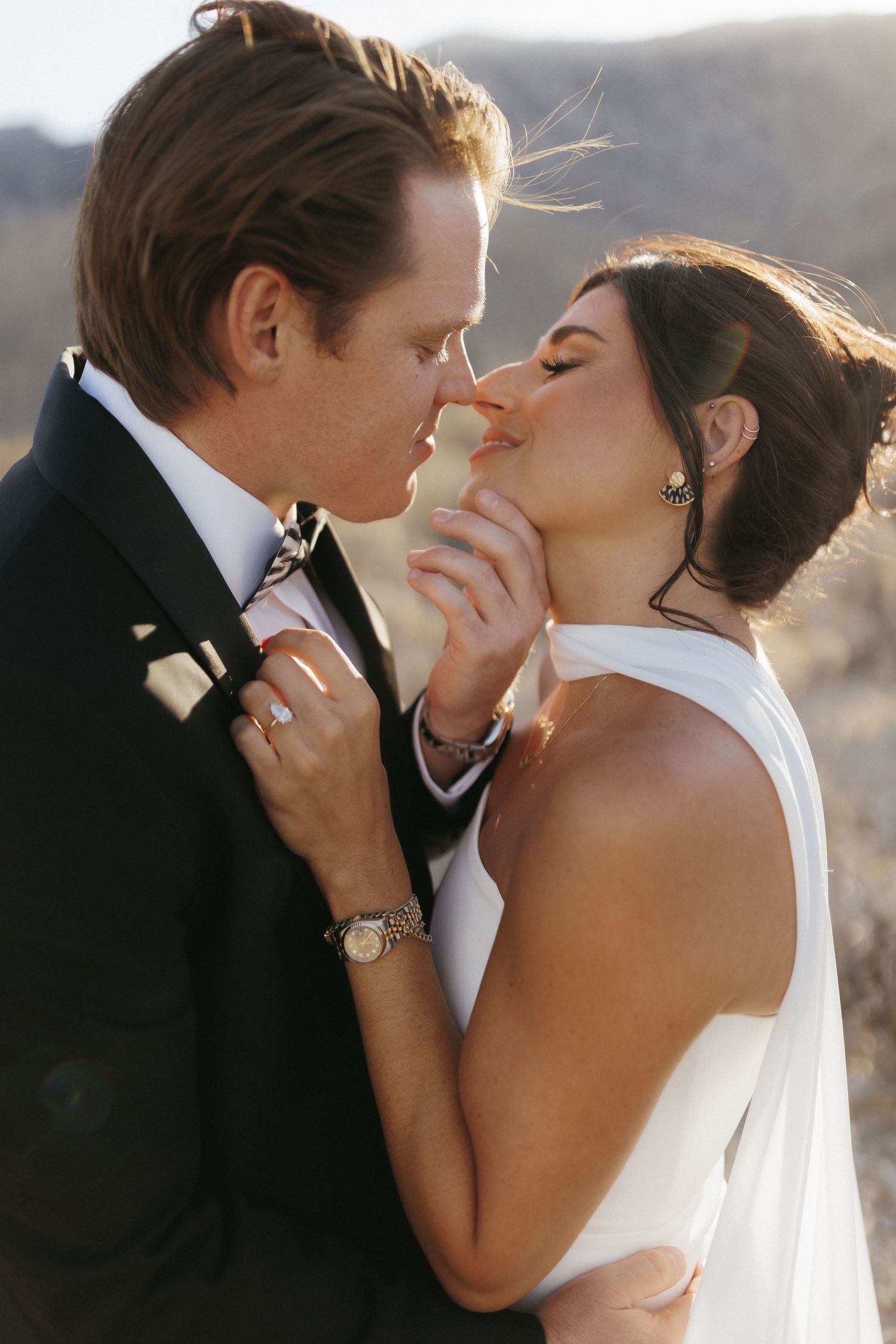 A bride and groom share a romantic kiss outdoors, with the bride in a white halter gown and groom in a black tuxedo.