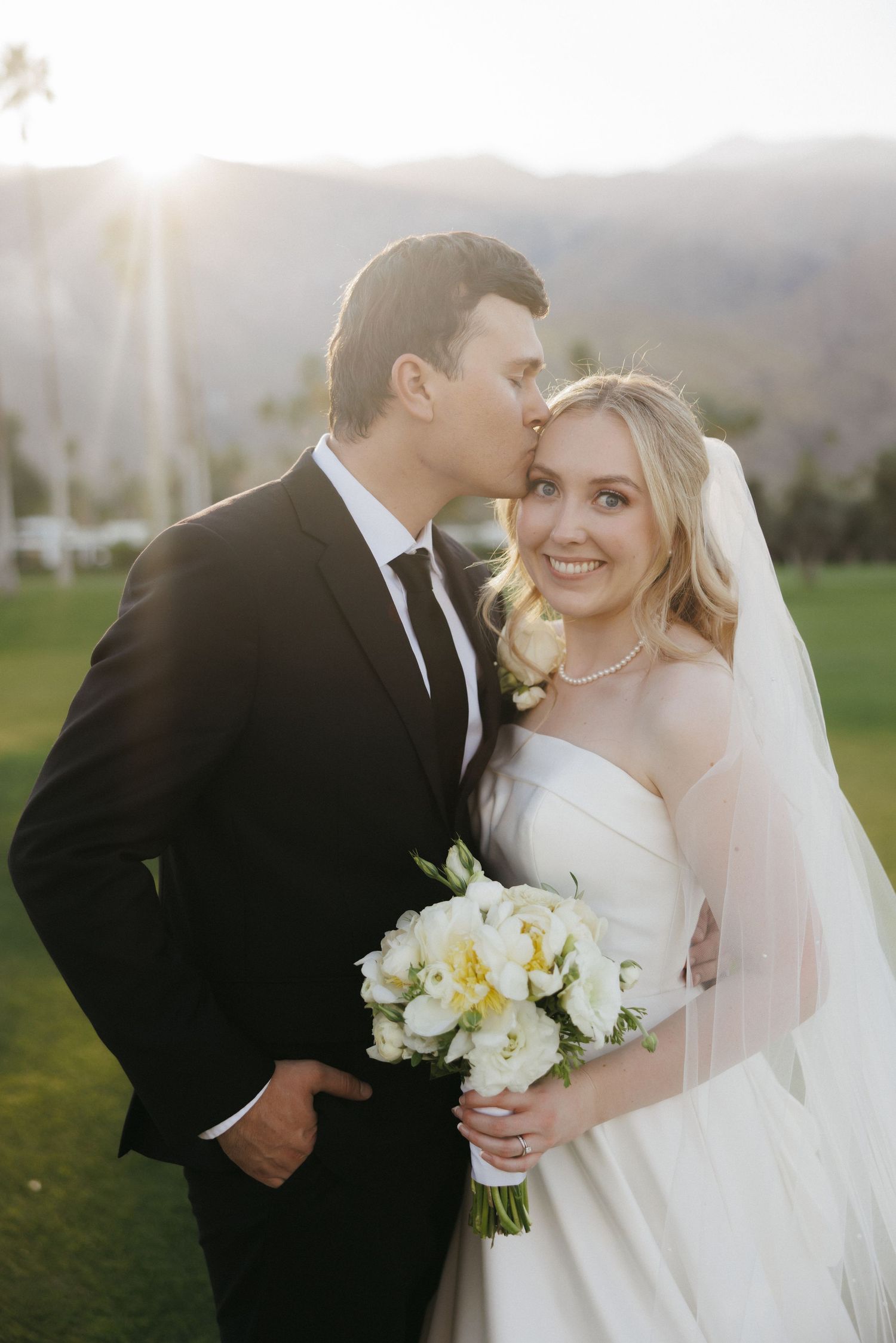 Groom kisses bride on cheek as she smiles holding white floral bouquet at sunset.