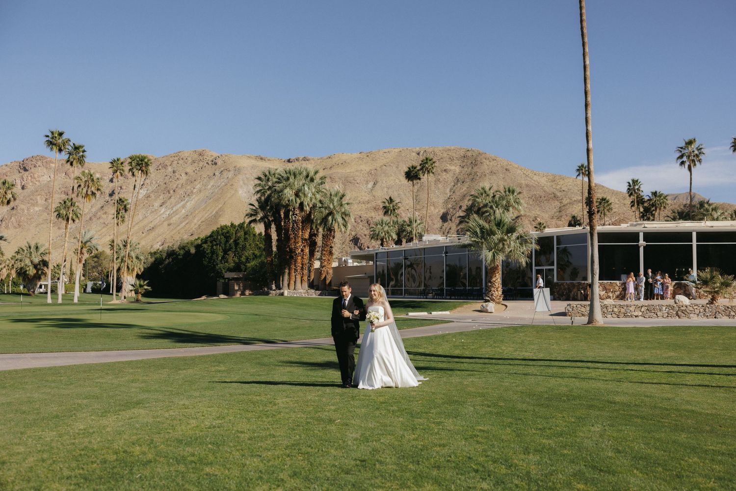Bride and groom portrait on lush green lawn with palm trees and mountains in Palm Springs.