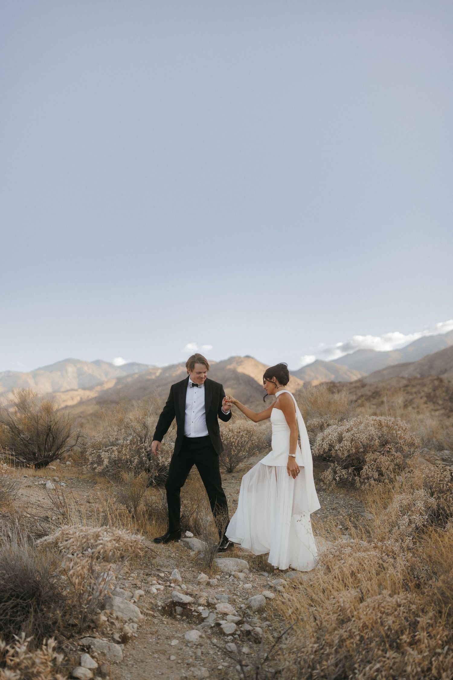 Groom helps bride in flowing white dress navigate a rocky desert trail with mountains in the background.
