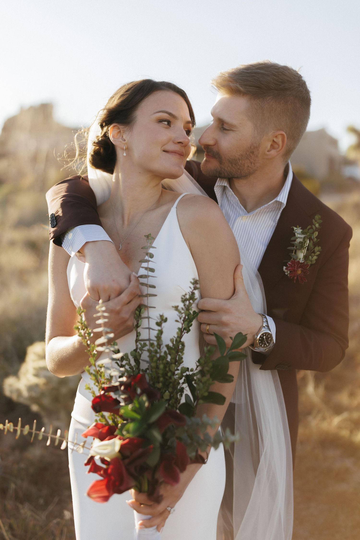 Groom in brown suit embraces bride holding red floral bouquet outdoors in golden sunlight.