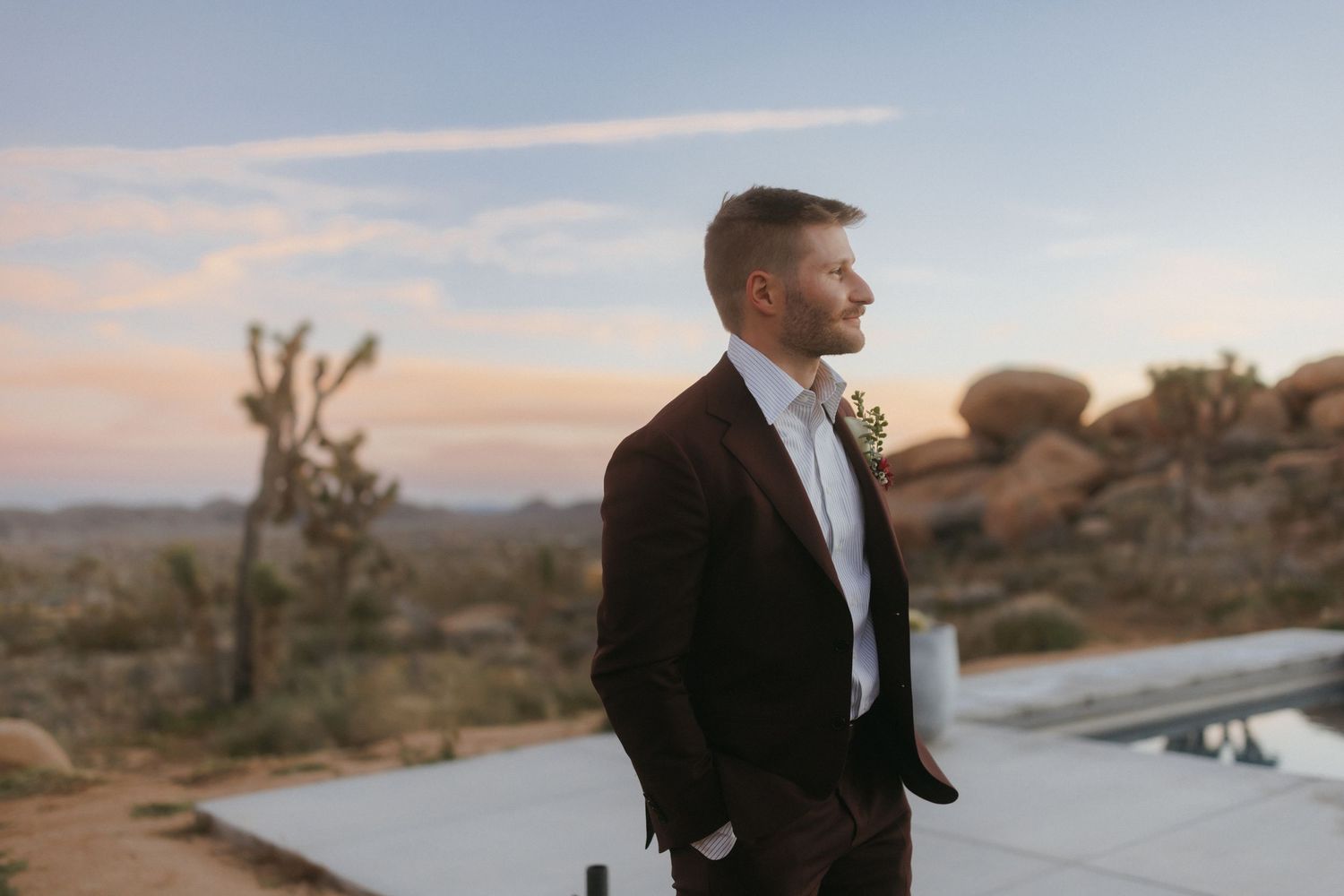 Dapper groom in dark suit stands by pool at desert sunset venue.
