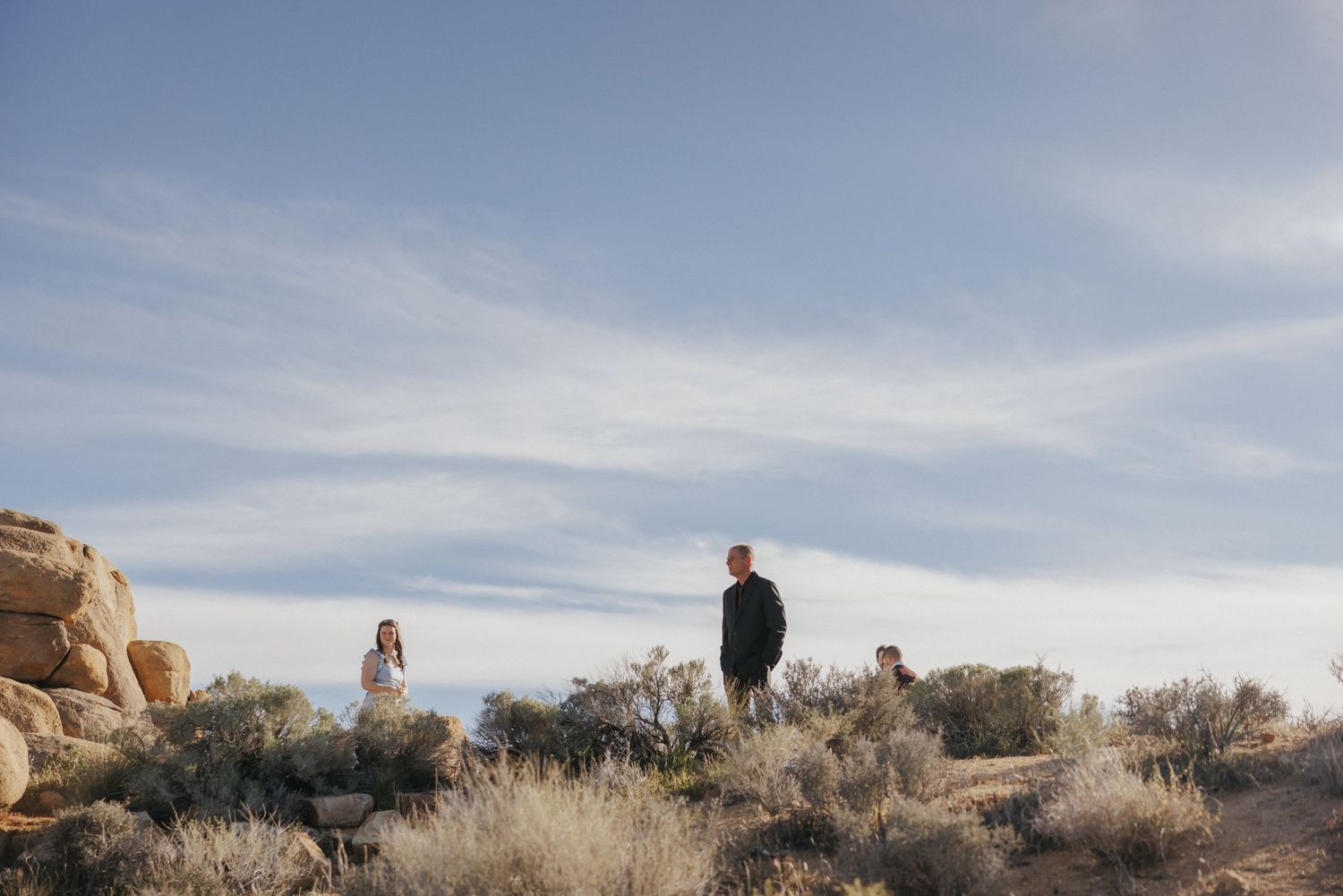 Three people stand among desert shrubs and rocky formations under a vast blue sky in an arid landscape.