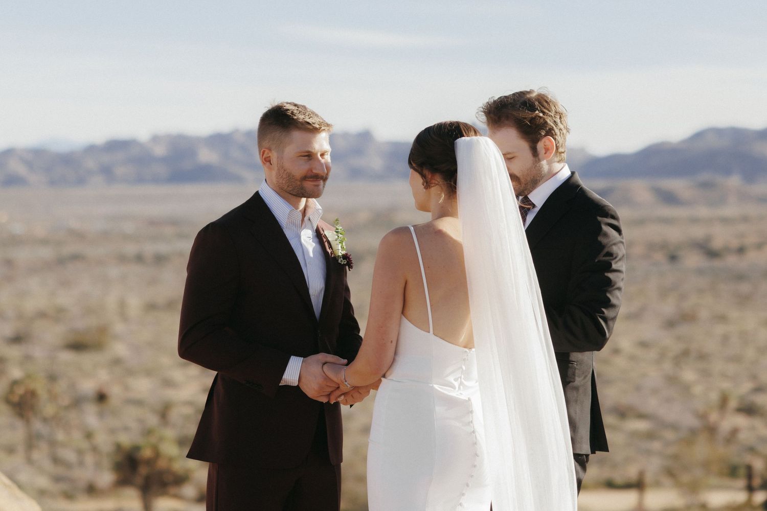 Bride and groom hold hands during outdoor desert wedding ceremony with mountain backdrop and officiant.