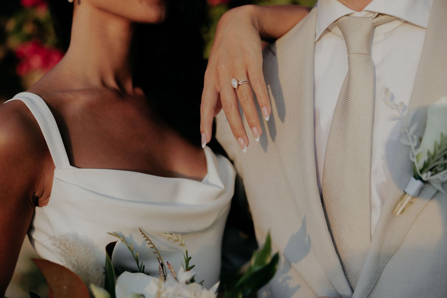 Close-up formal wedding photo showing white wedding dress strap and beige suit with floral boutonniere against blurred background.