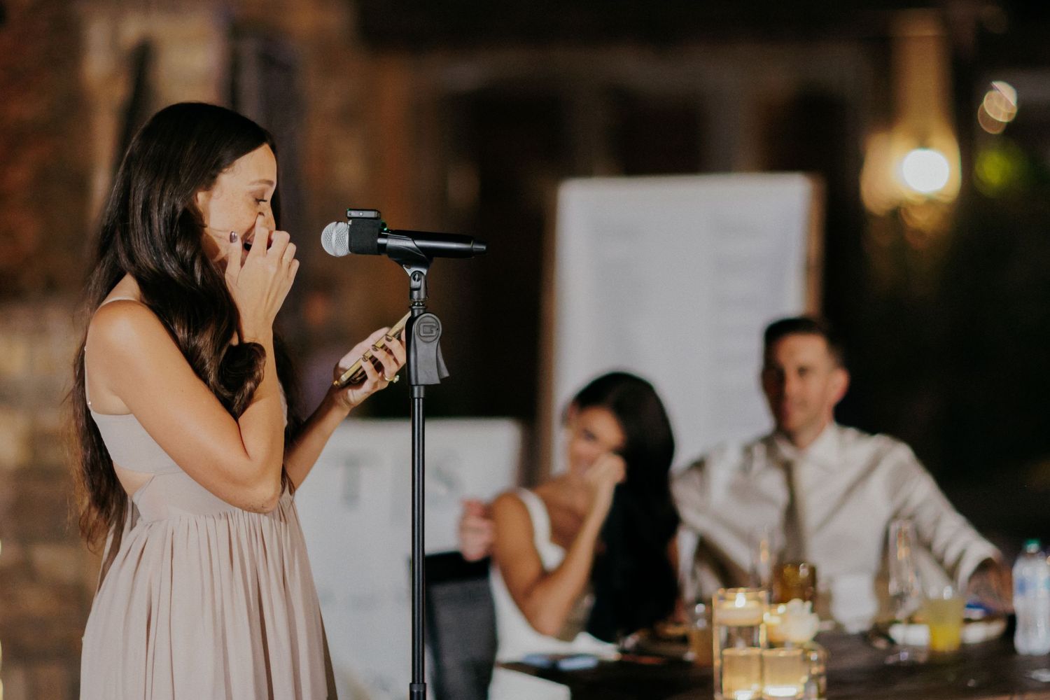 Someone delivers a heartfelt speech at a wedding reception while guests listen attentively in a dimly lit venue.