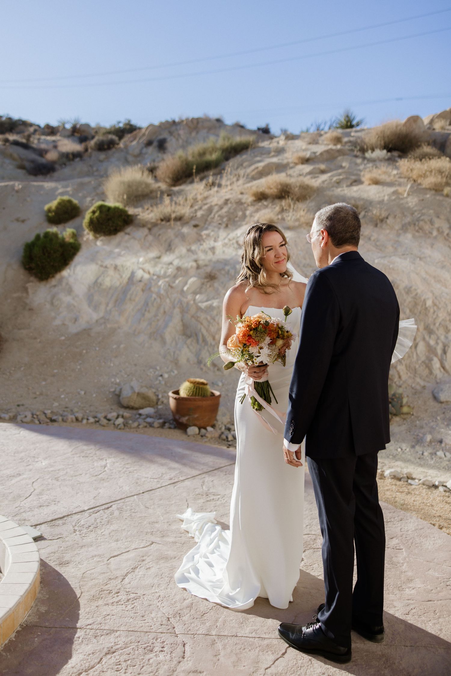 A couple walking along a desert cliff path on their wedding day with a white dress and black tuxedo in view.