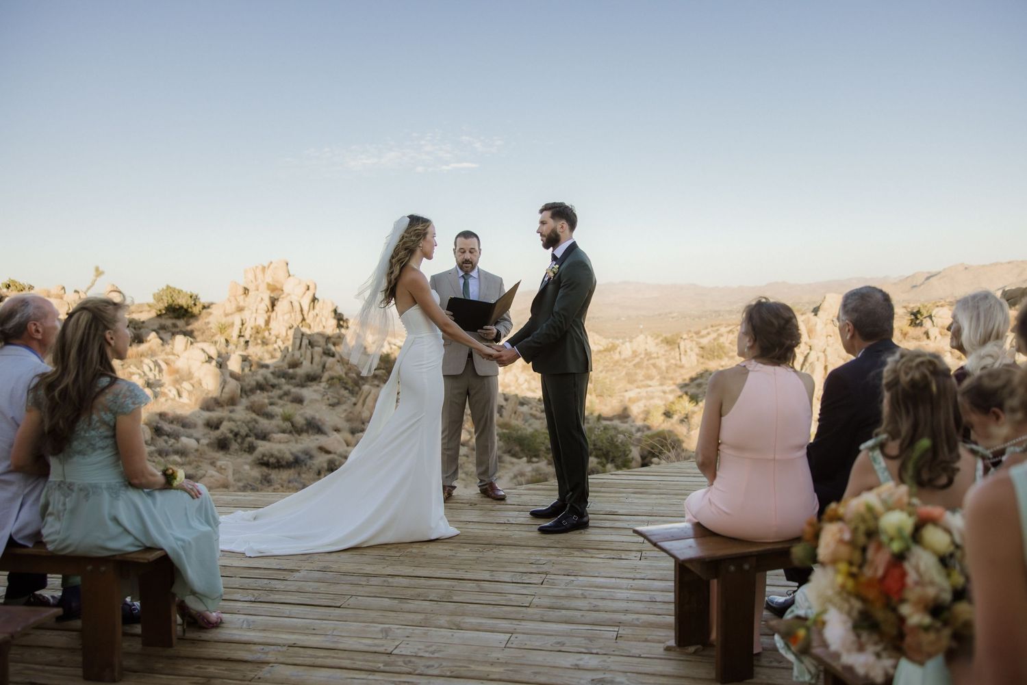 Ceremony participants gather for an outdoor desert wedding at sunset with mountains and rock formations in the background.