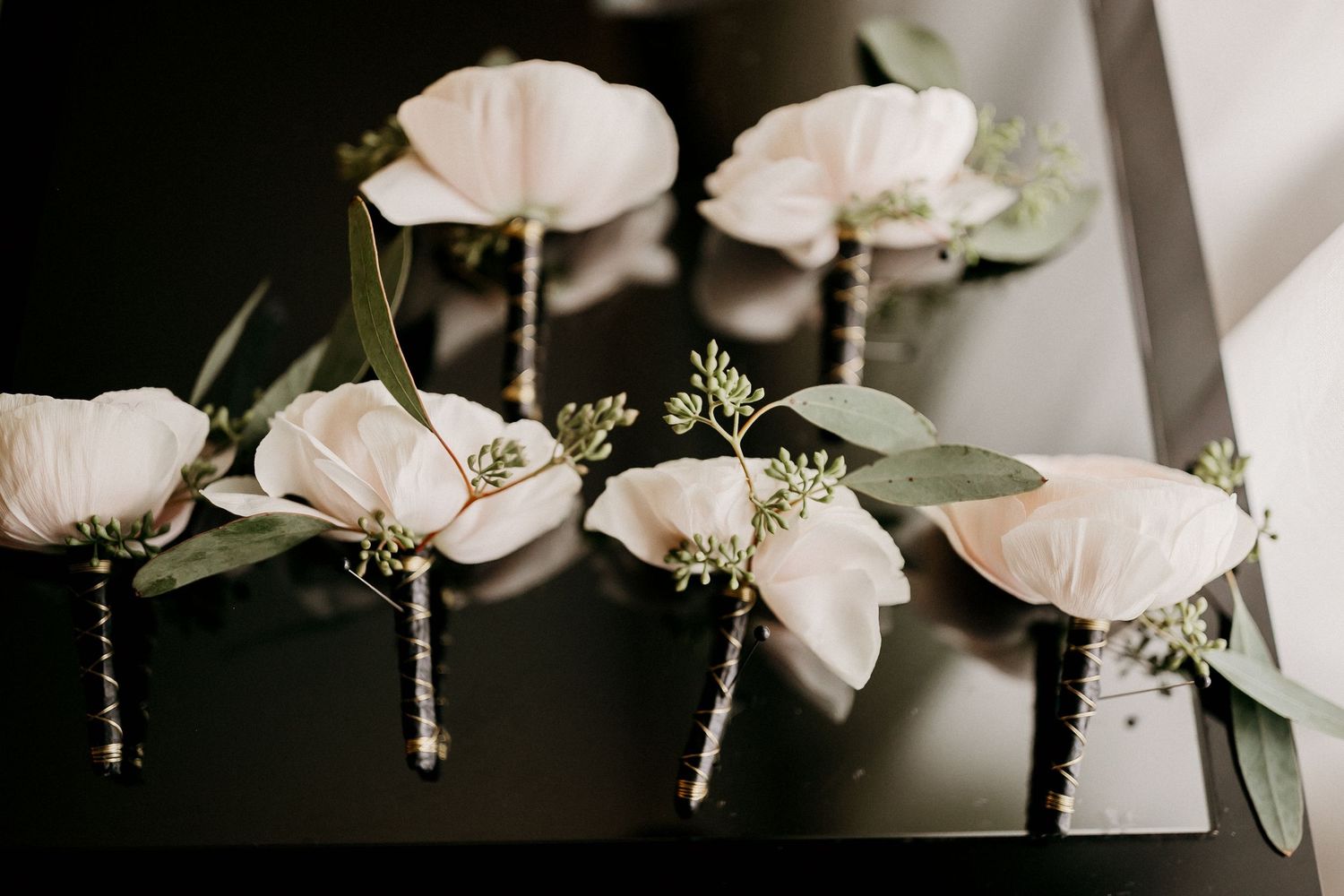 Row of elegant white rose boutonnieres with eucalyptus leaves lined up against dark background.
