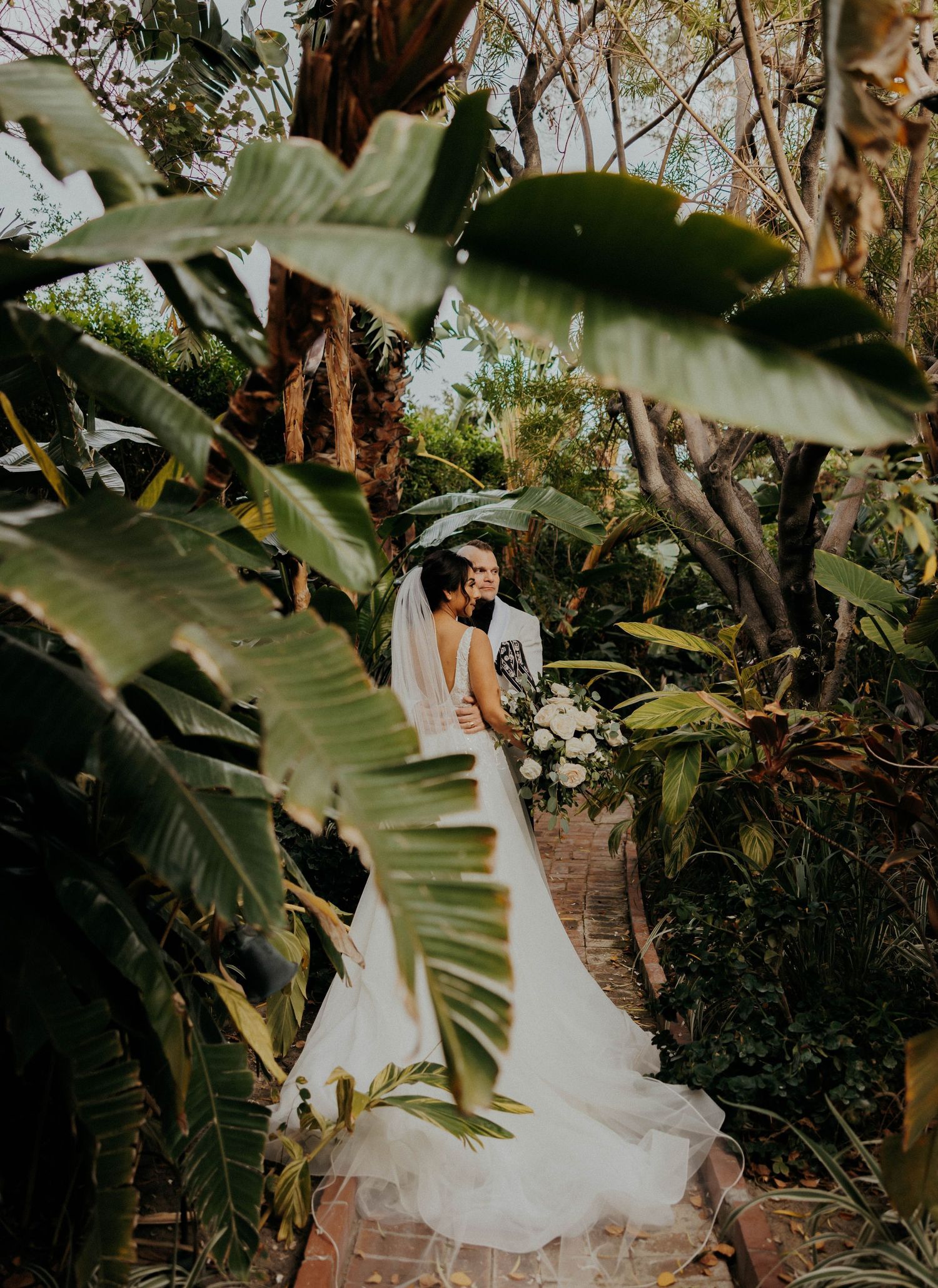 A romantic wedding moment framed by large tropical leaves and greenery along a brick pathway.