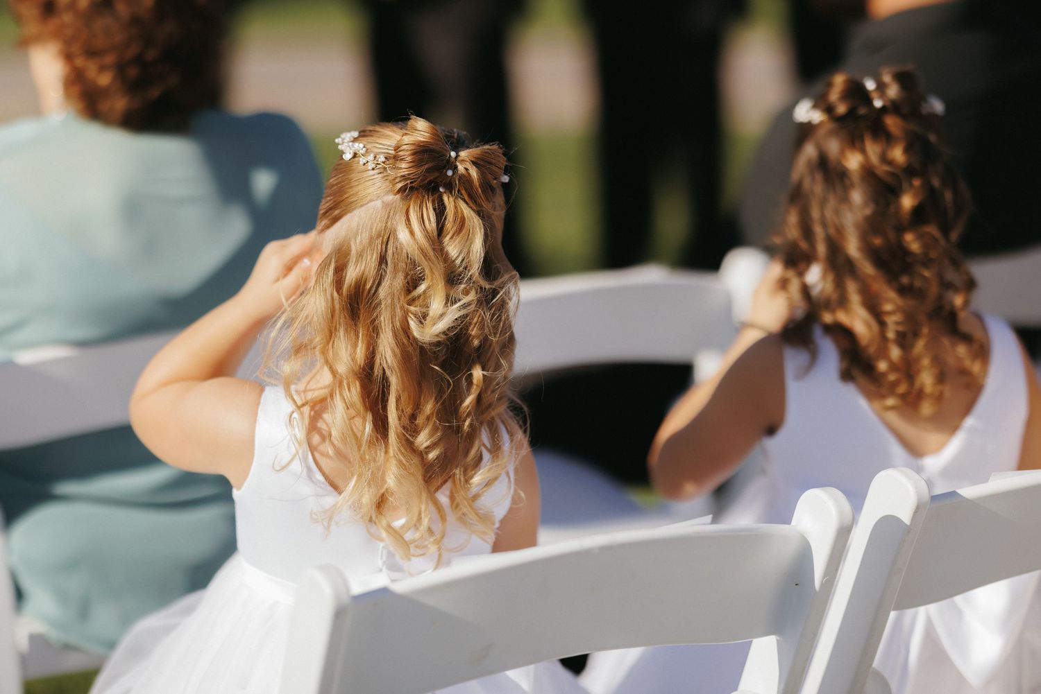 Two flower girls with elegant hairstyles seated in white chairs at an outdoor wedding ceremony.