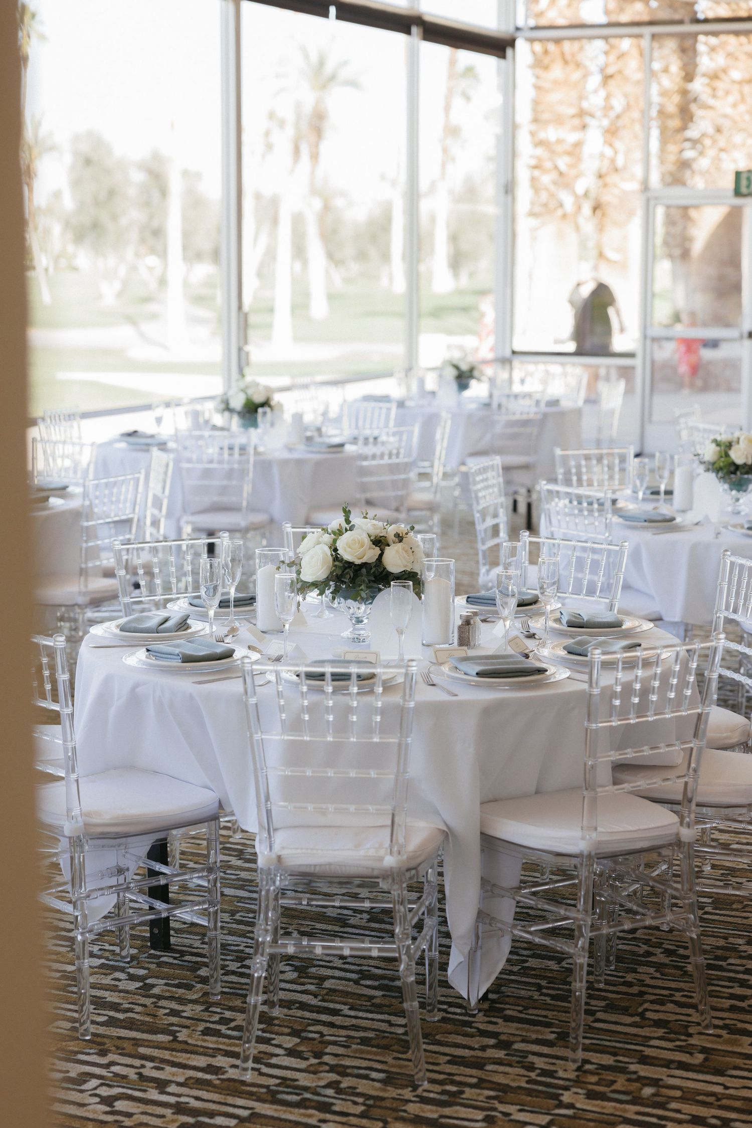 White wedding reception tables with clear chiavari chairs and white rose centerpieces near large windows with garden views.