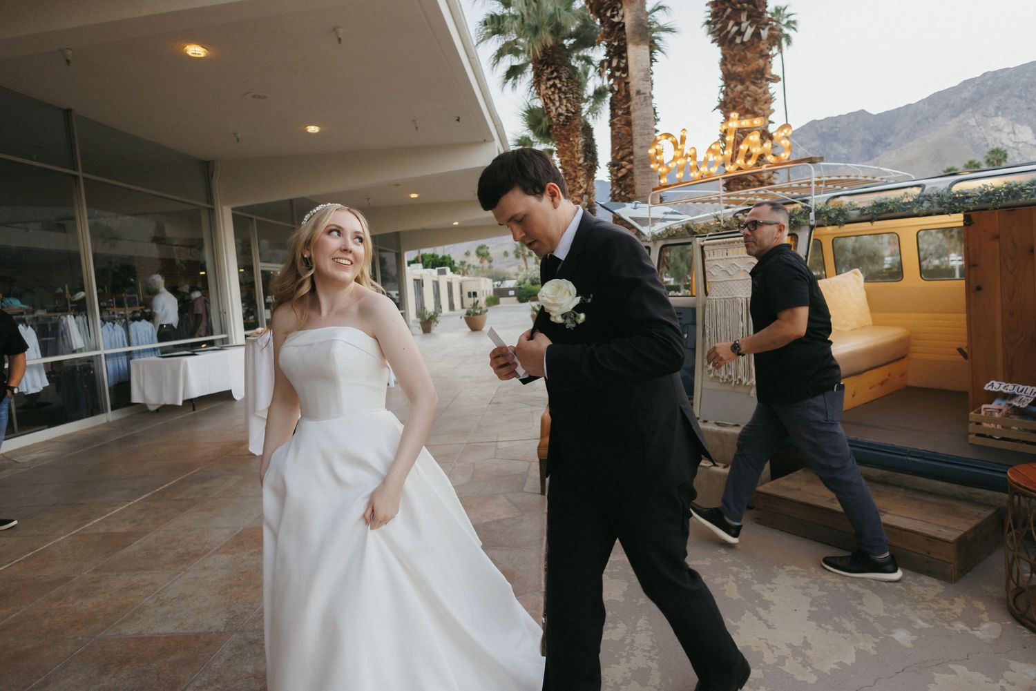 Smiling bride in white strapless gown walking with groom in black tuxedo at outdoor wedding.