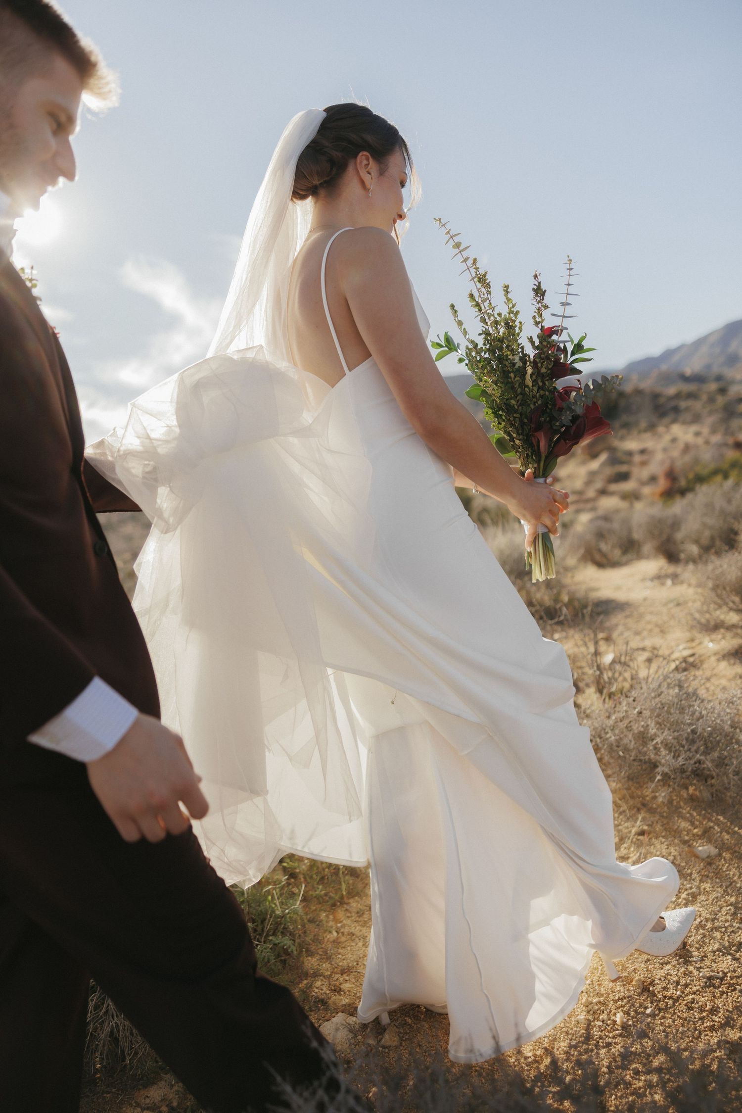 Bride in flowing white gown with veil holds bouquet while walking hand-in-hand with groom in desert landscape.