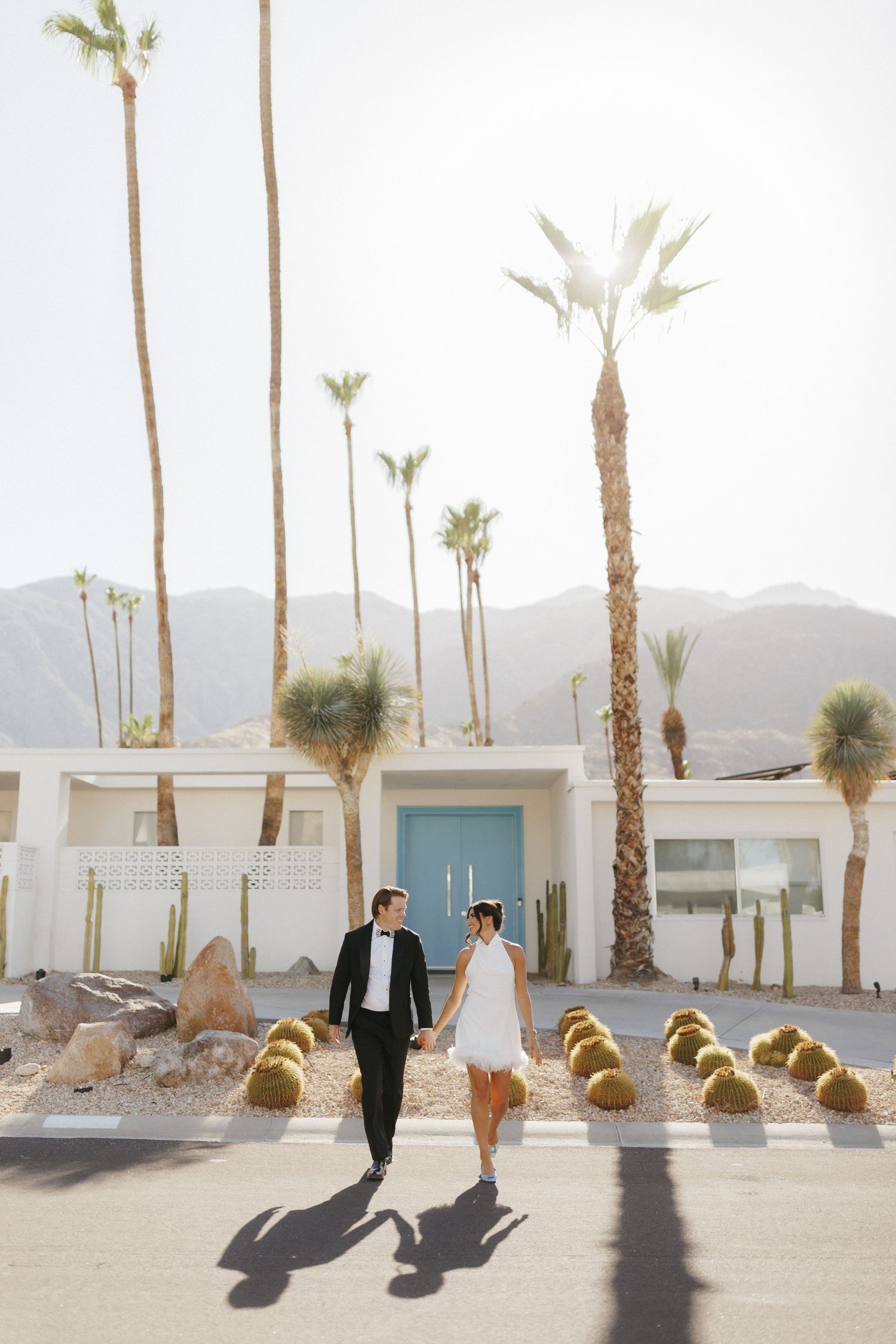 Couple holding hands walking in front of a mid-century modern home with palm trees and mountains in Palm Springs.