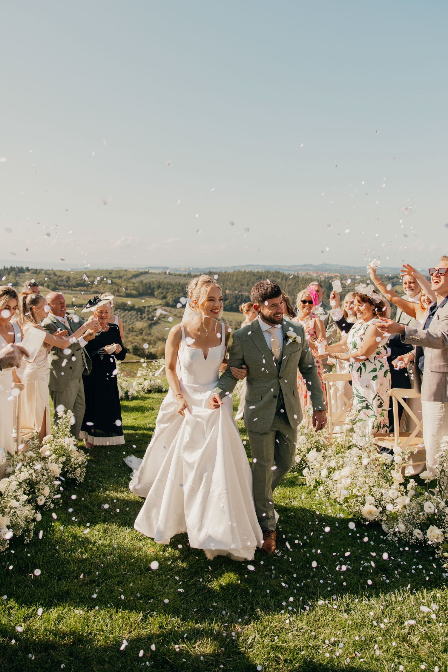 Wedding guests toss petals as couple walks down aisle in outdoor ceremony with scenic backdrop.