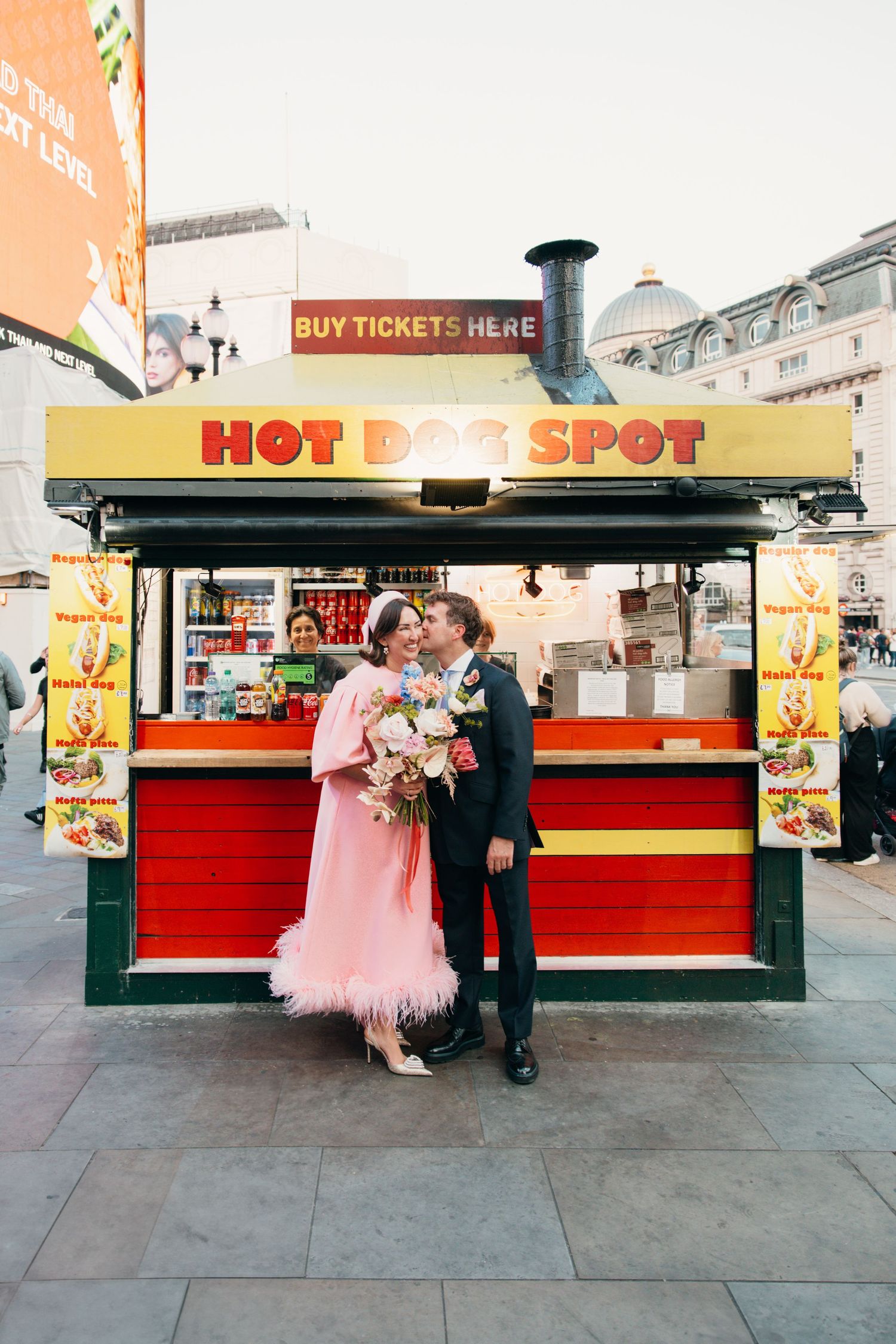 Newlyweds share romantic moment in front of red and yellow hot dog stand.