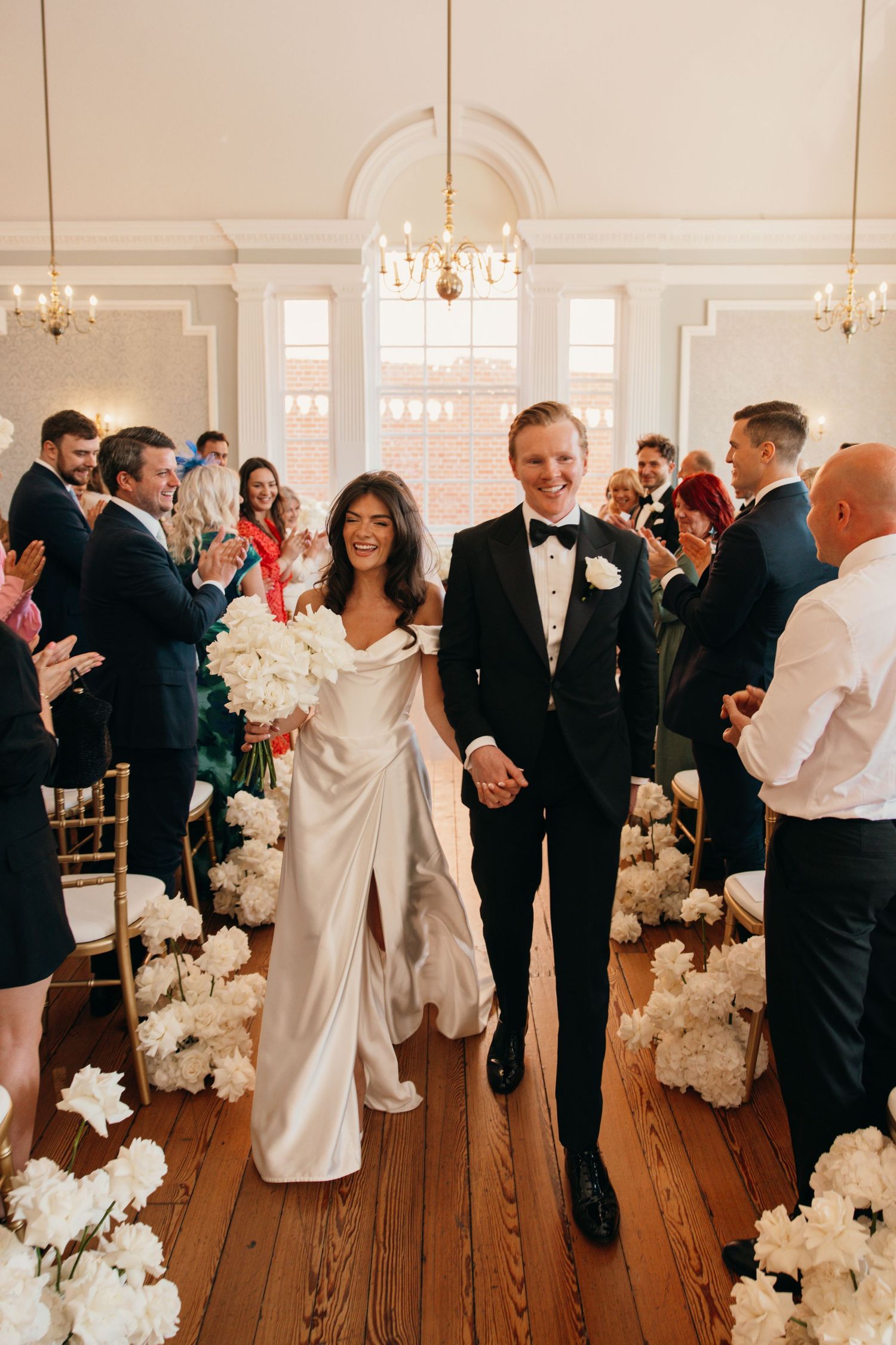 Bride and groom walk down the aisle holding hands, surrounded by white floral arrangements and applauding guests.