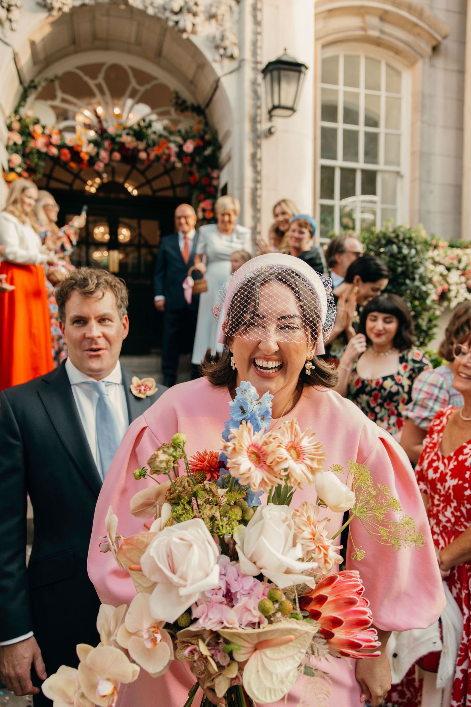 Wedding guests celebrate as couple exits ornate building entrance decorated with floral arrangements.