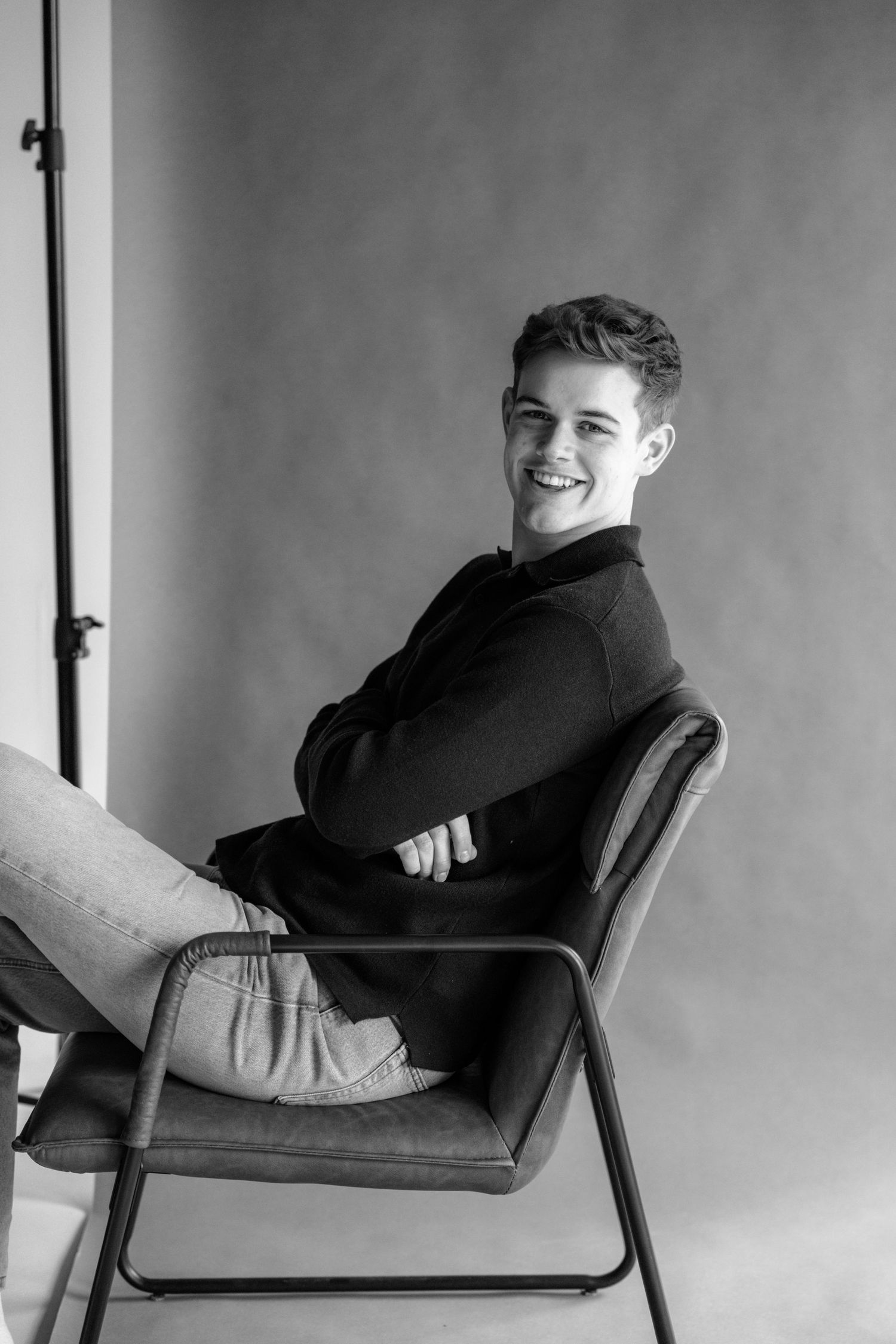 Black and white portrait of person sitting casually on chair with joyful expression.