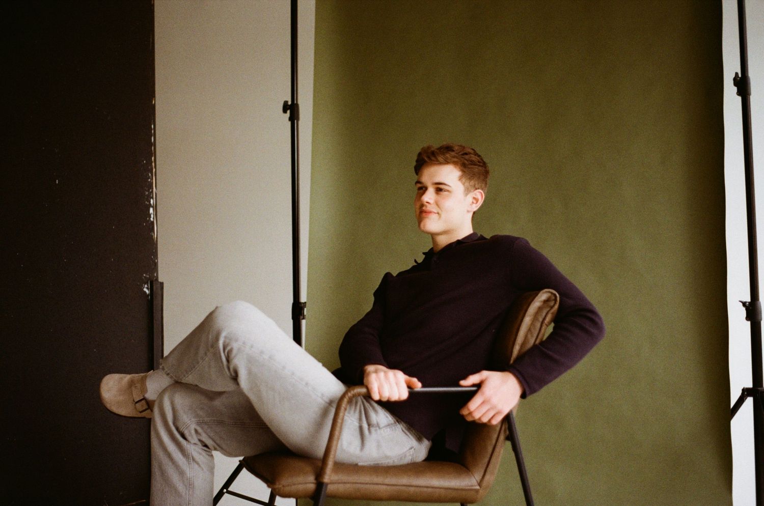A person sits casually in a folding chair wearing a black sweater and light colored pants against a dark backdrop.