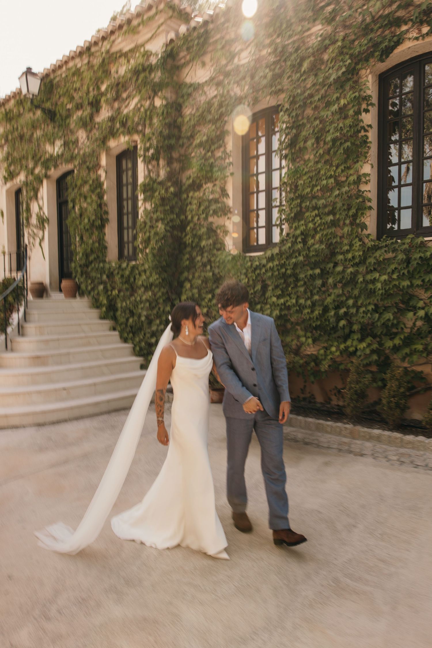 Bride and groom walking hand in hand outside an ivy-covered stone villa during golden hour.