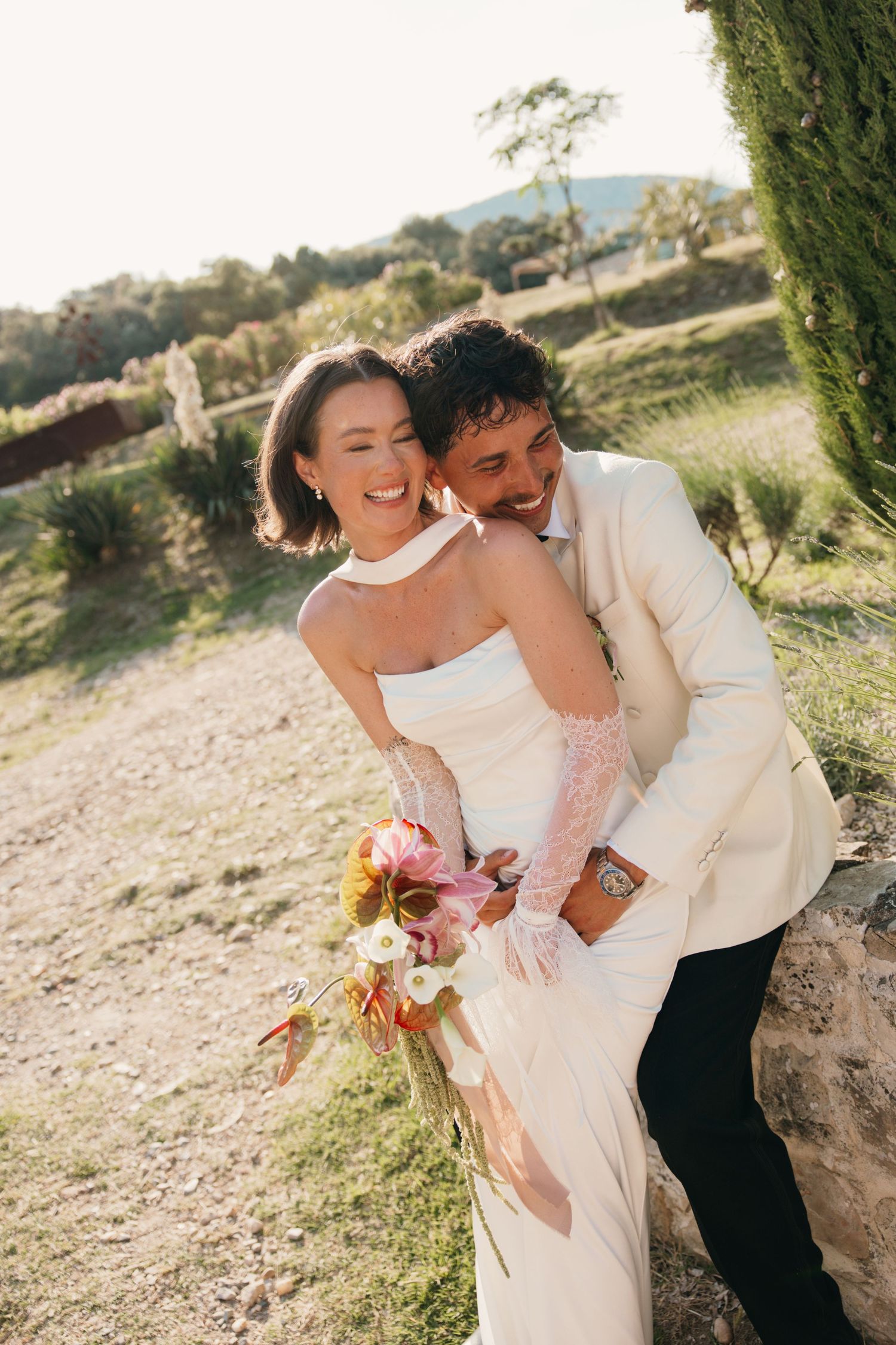 A joyful bride in a white strapless gown holds a bouquet while a groom in a cream suit embraces her outdoors.