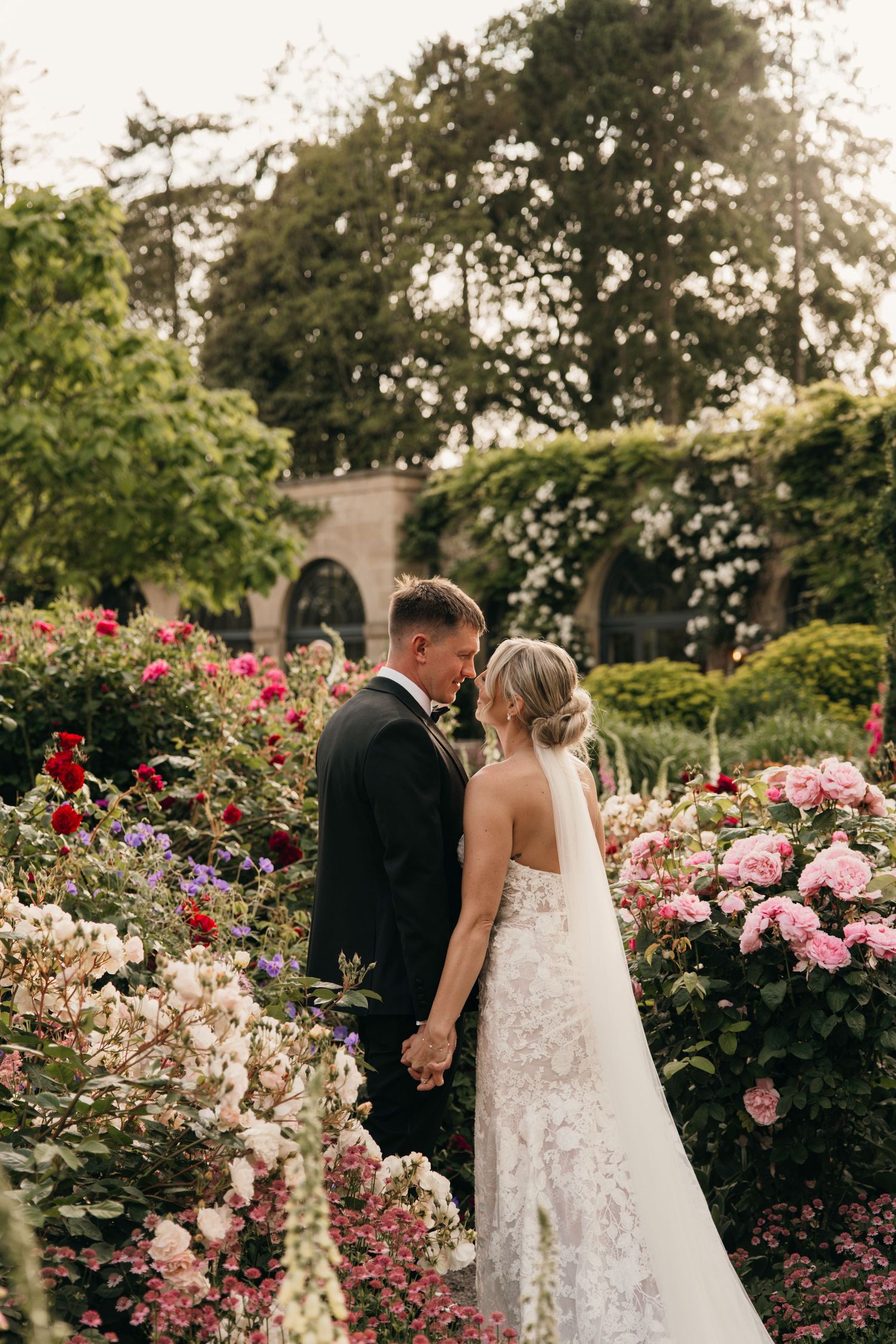 A couple shares an intimate moment among blooming pink roses and flowers in a lush garden setting on their wedding day.
