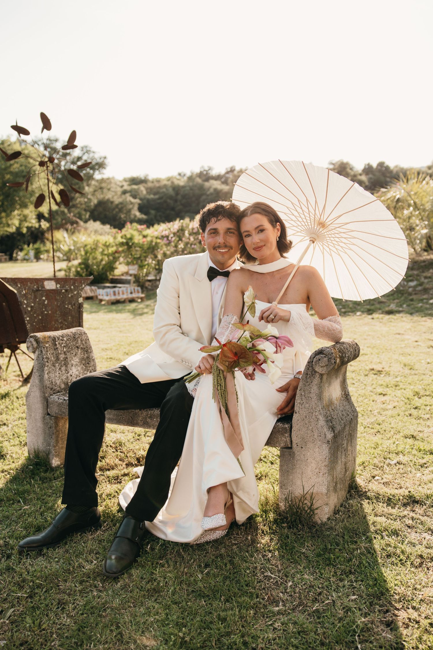 Bride and groom sitting on stone bench outdoors, bride holding parasol and floral bouquet, groom in white tuxedo jacket.