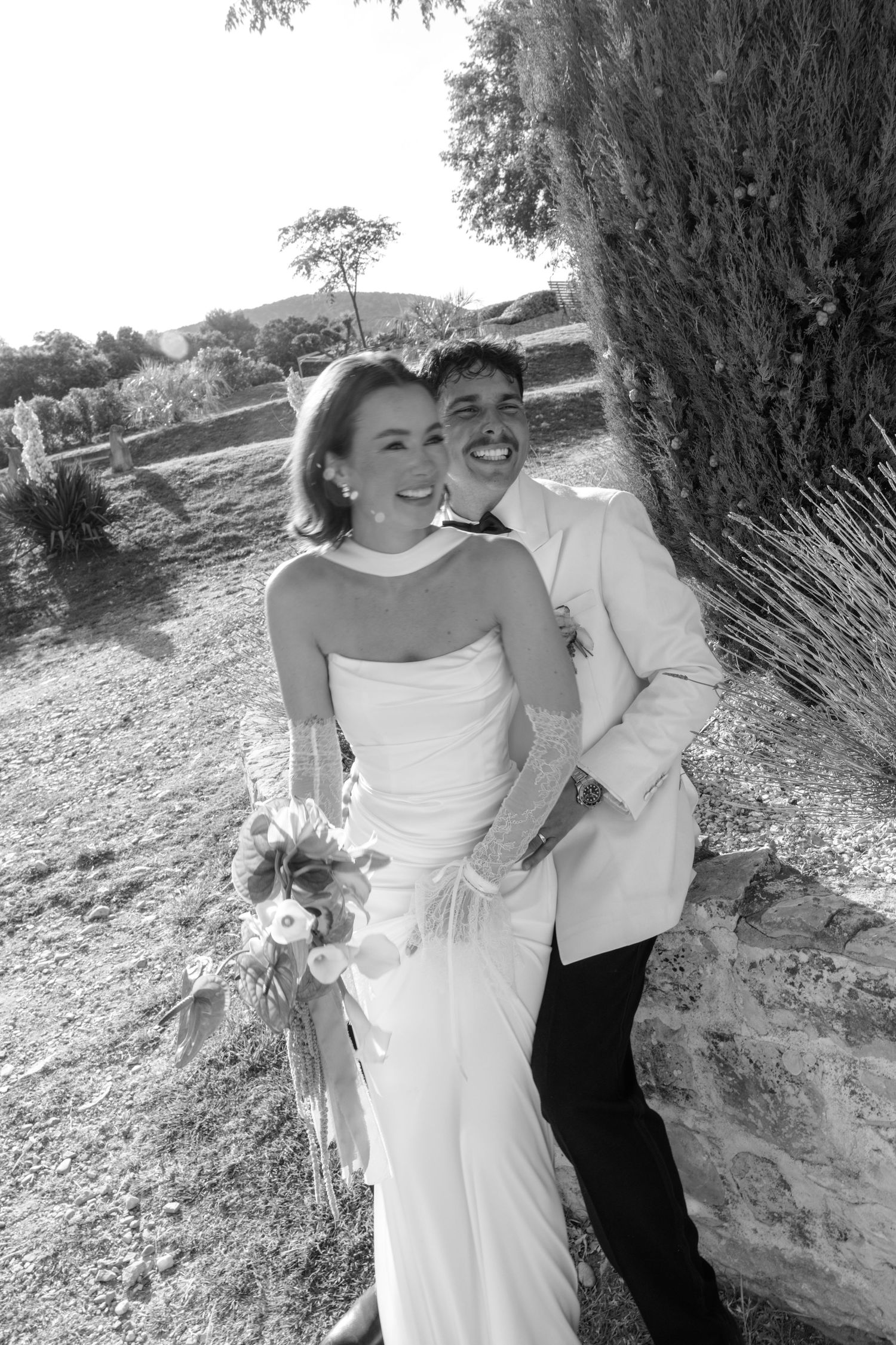 Black and white photo of a smiling bride in strapless gown and groom in light blazer posing outdoors on their wedding day.