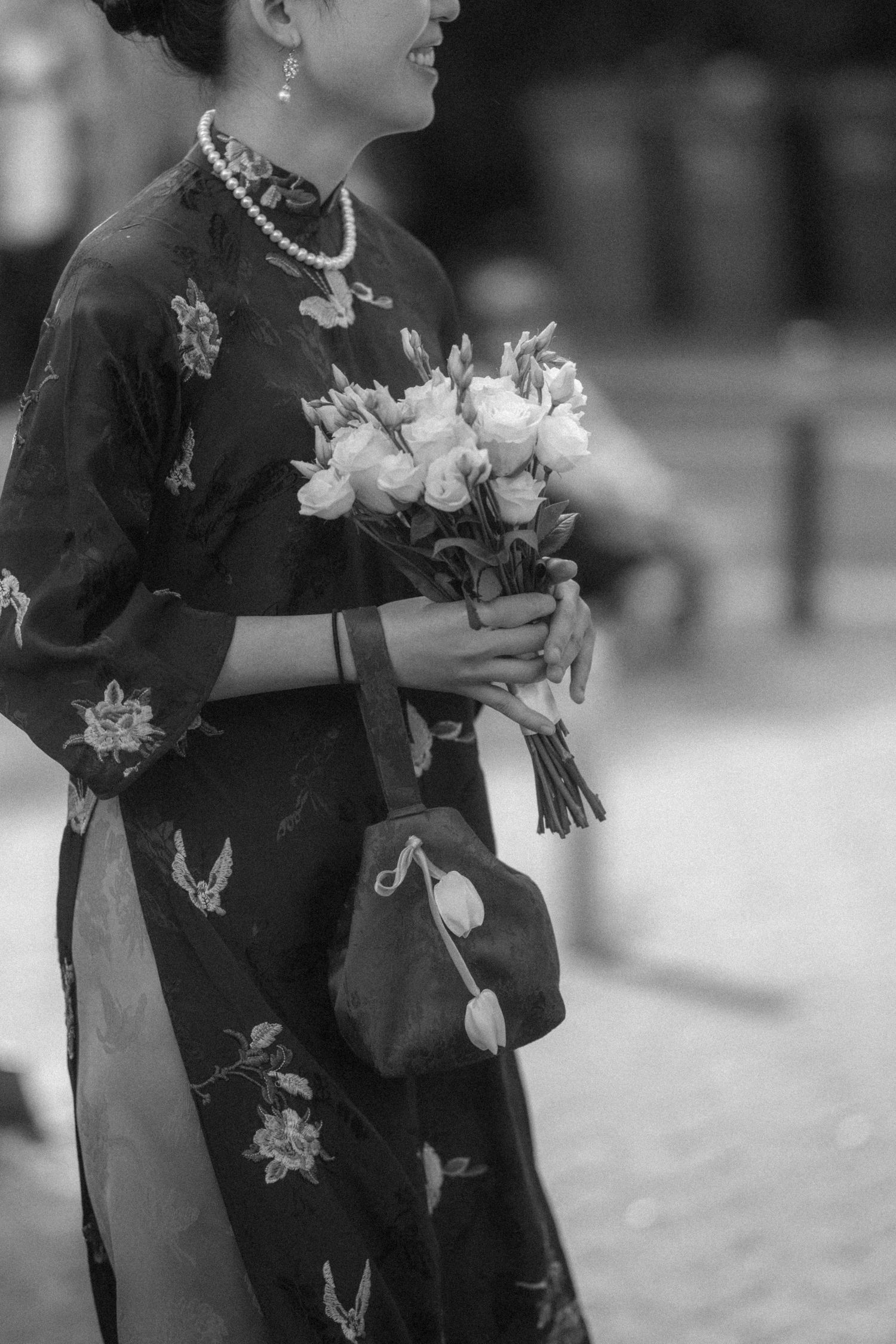 Black and white photo of woman in floral qipao holding rose bouquet with pearl necklace and small handbag.