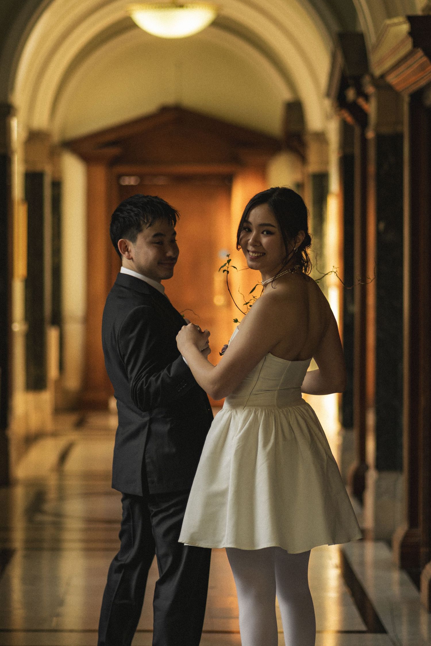 Couple in elegant attire pose in ornate hallway, woman in white mini dress smiles over shoulder, man in black suit.