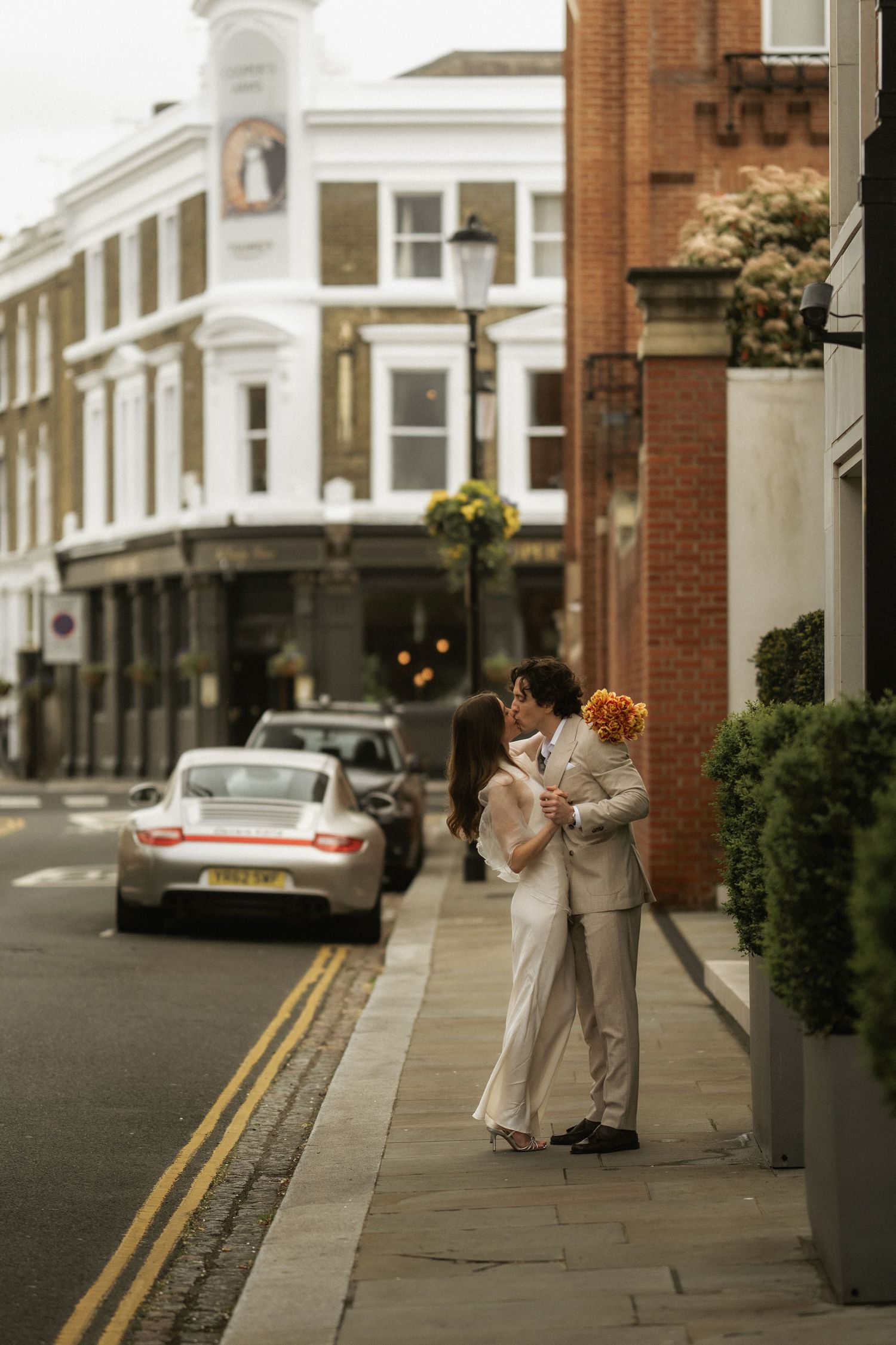 Couple sharing a kiss on a London street corner, bride holding orange bouquet, groom in beige suit, classic Porsche in background.
