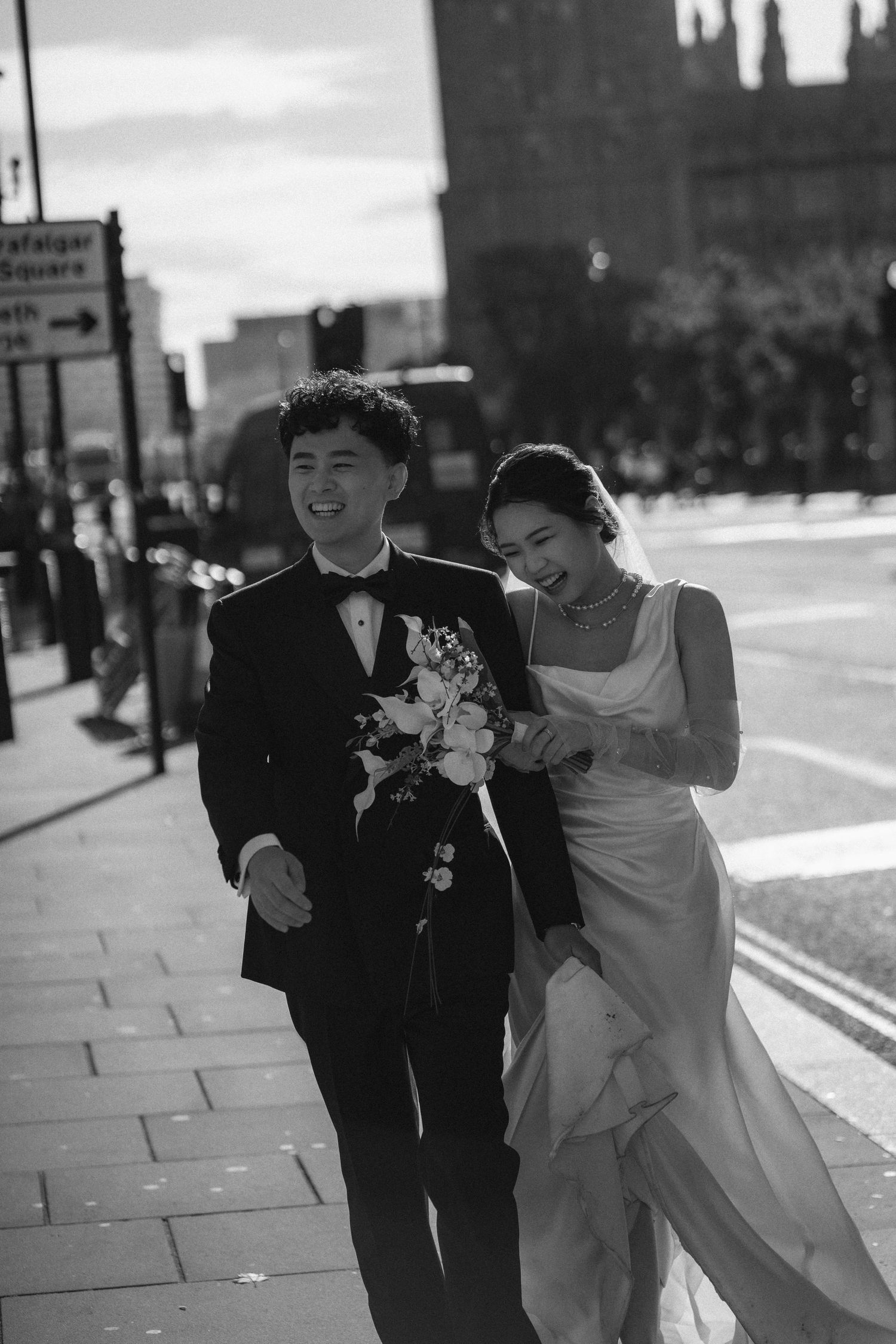 Black and white photo of a smiling bride and groom walking together on a London street, bride holding a bouquet.