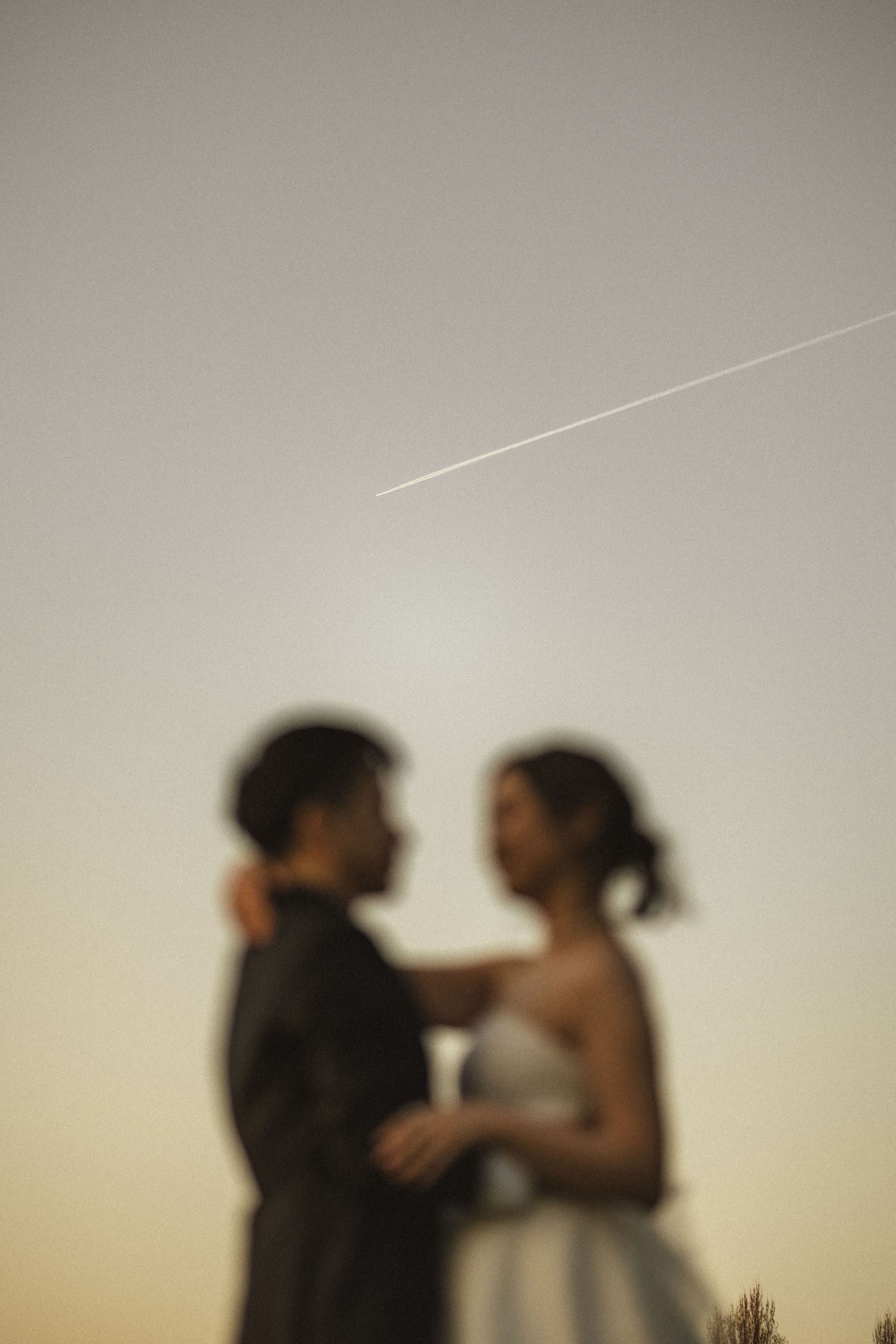 A blurred silhouette of a couple in wedding attire embracing at sunset with a contrail in the sky above.