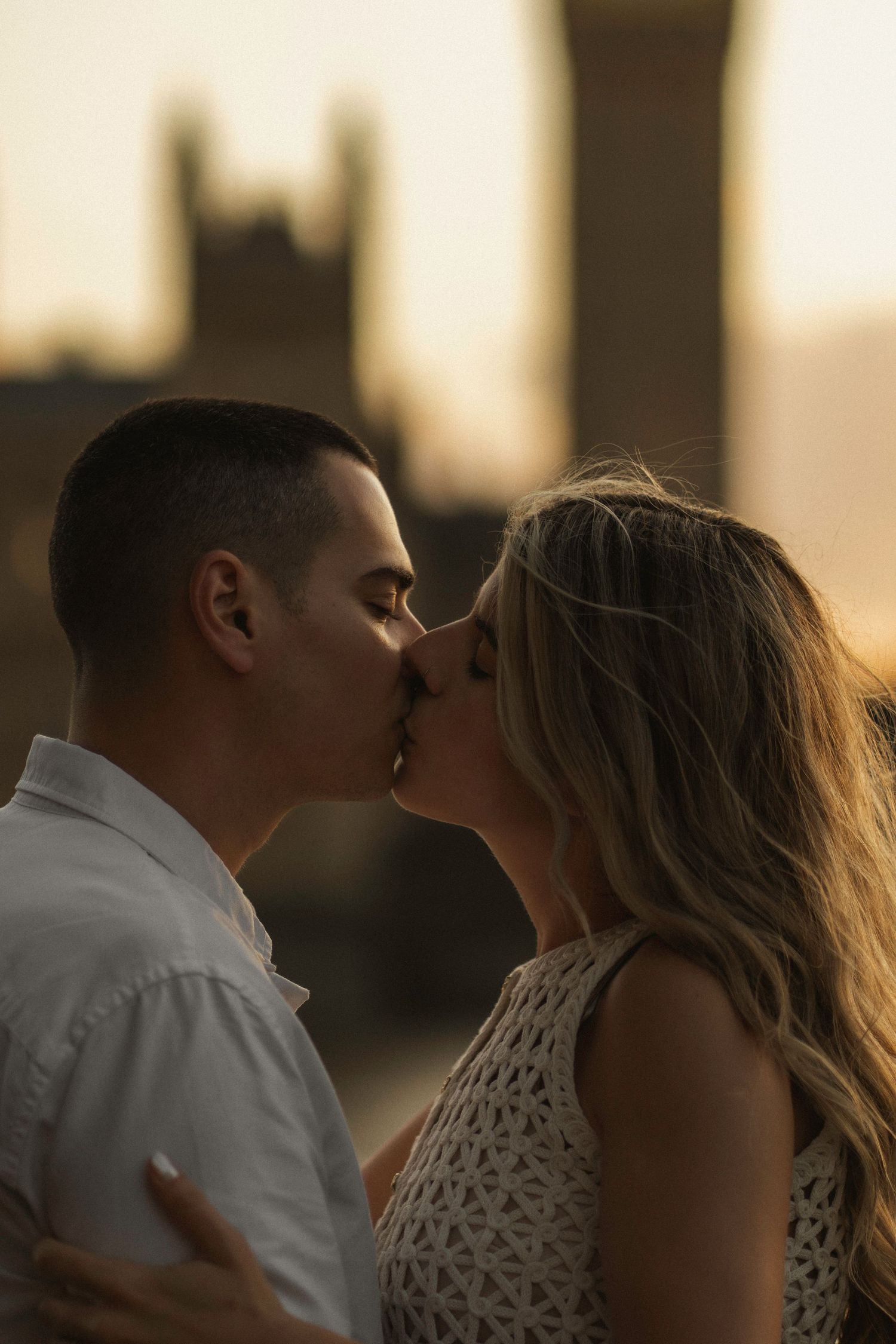 A romantic couple shares a tender kiss at sunset with a silhouetted building in the background.