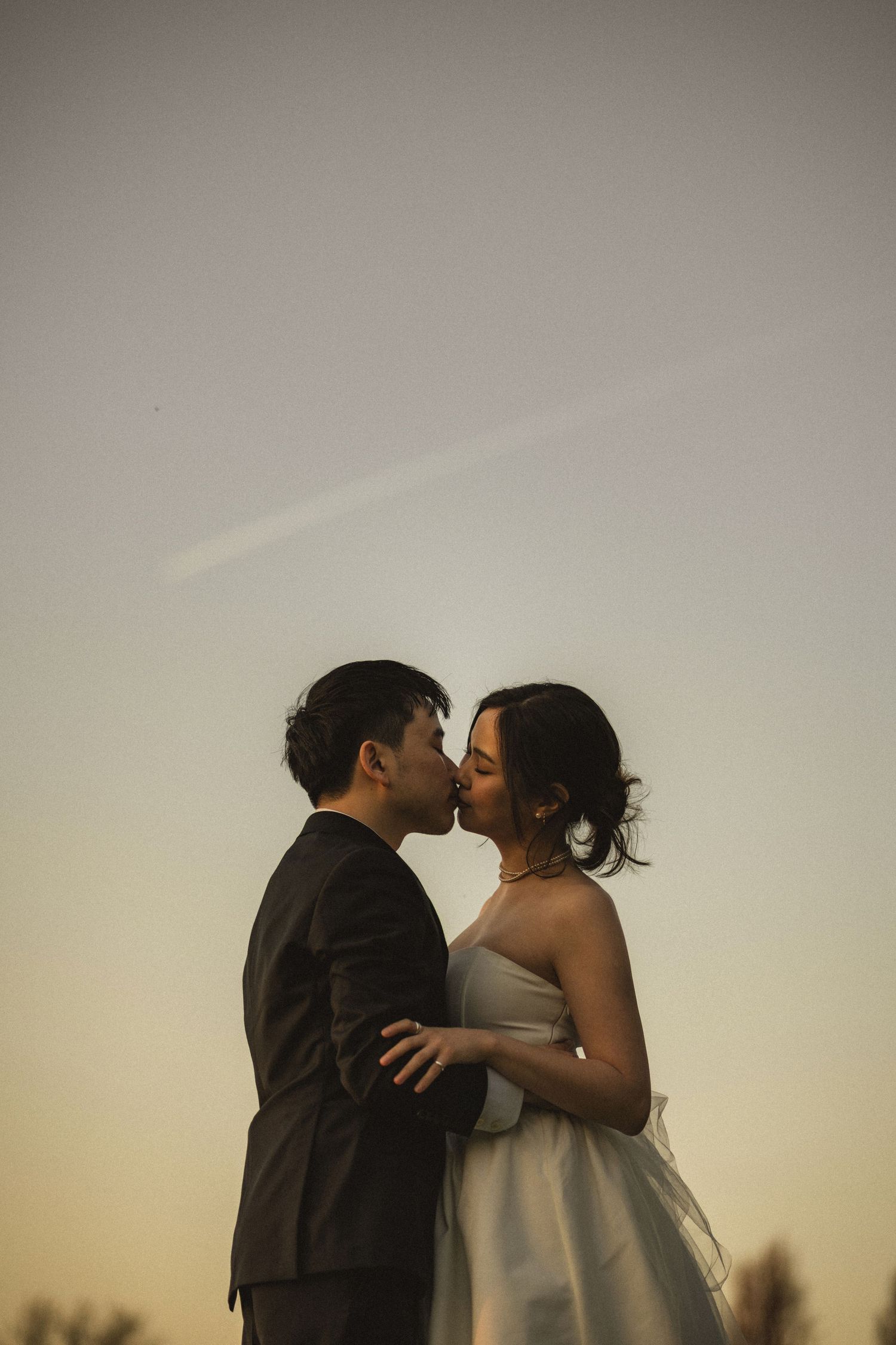 A couple shares a romantic kiss at sunset, the bride in a white gown and groom in a dark suit, silhouetted against a warm sky.