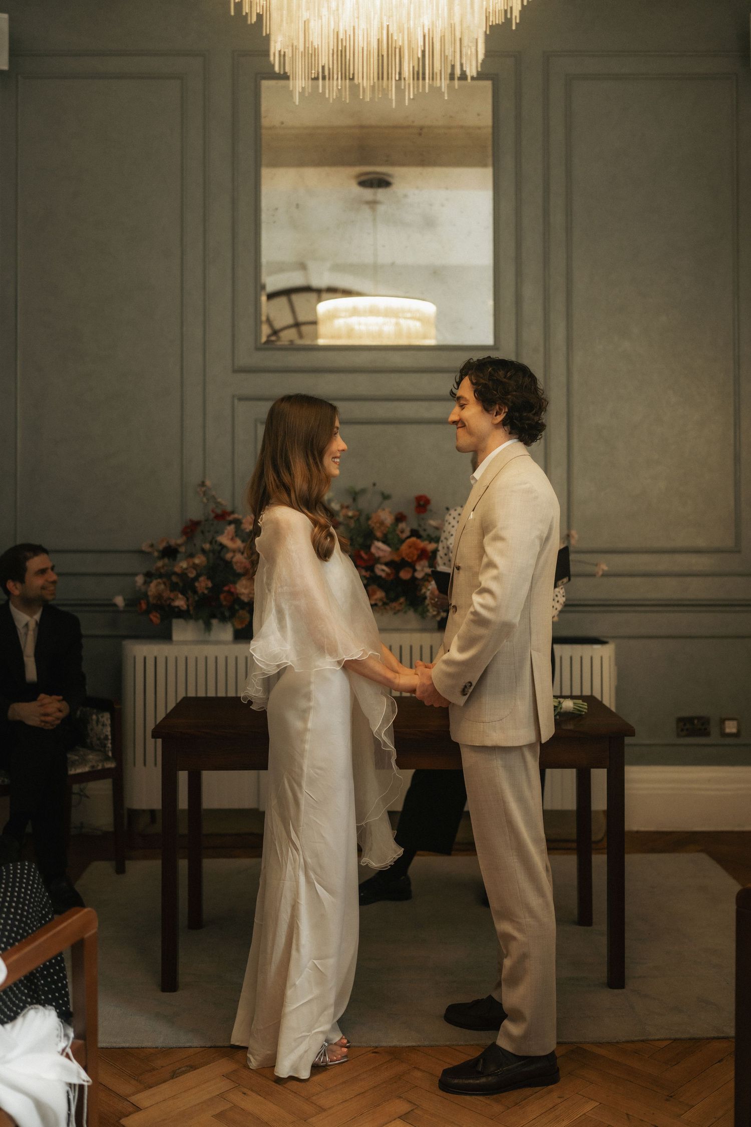 Couple holding hands at elegant wedding ceremony in a grey-walled room with chandelier and floral arrangement.
