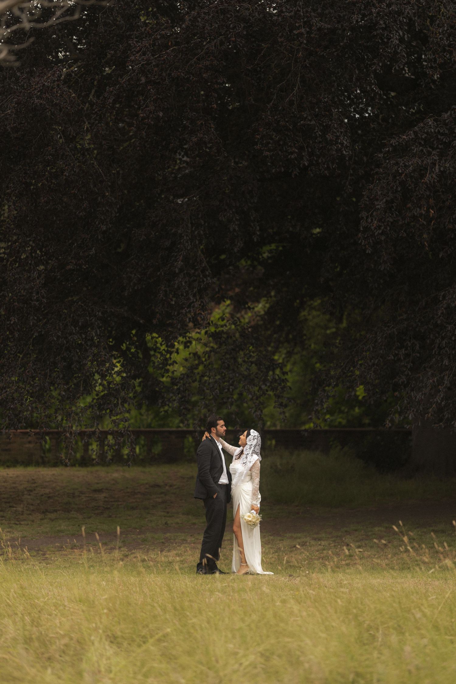 A bride and groom share a romantic moment in a sunlit meadow surrounded by lush dark trees on their wedding day.