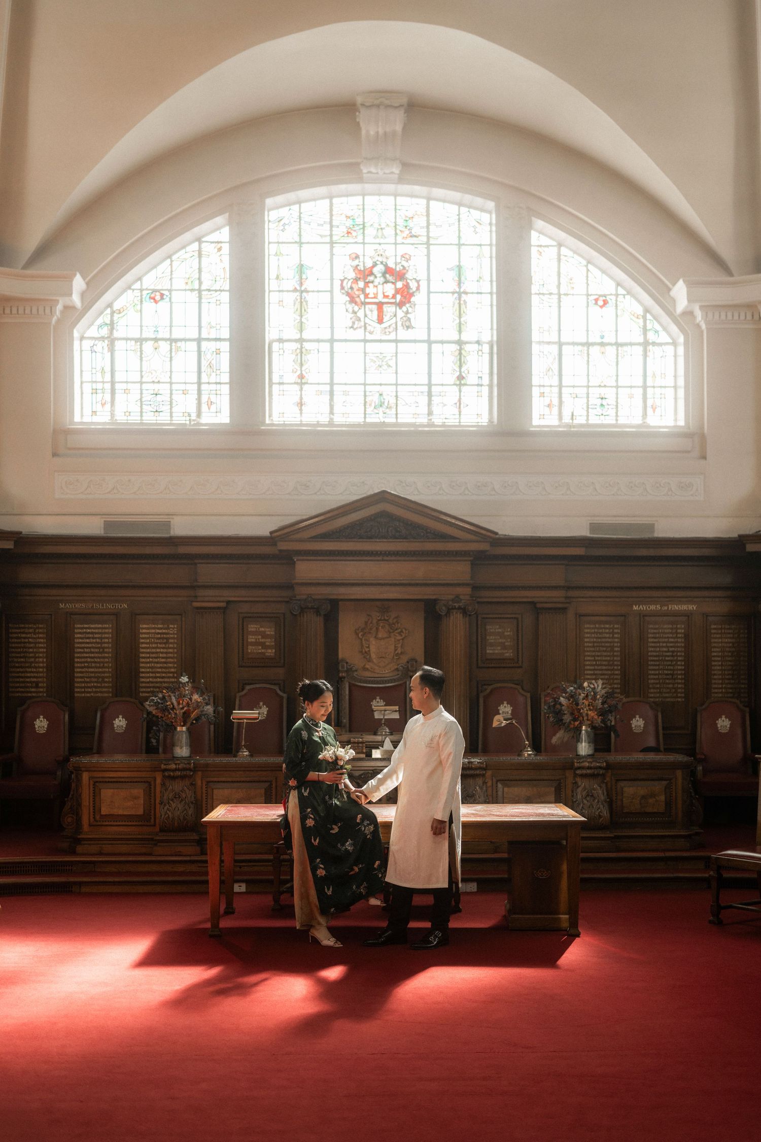 Couple in traditional Vietnamese ao dai attire standing at altar in elegant church with stained glass windows.