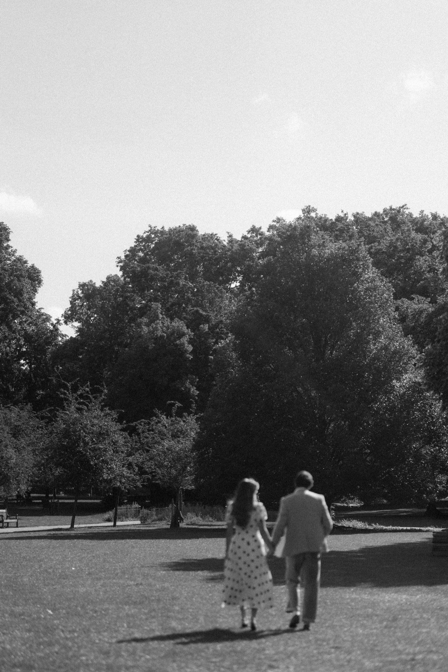 Black and white photo of a couple holding hands walking through a lush green park on a sunny day.