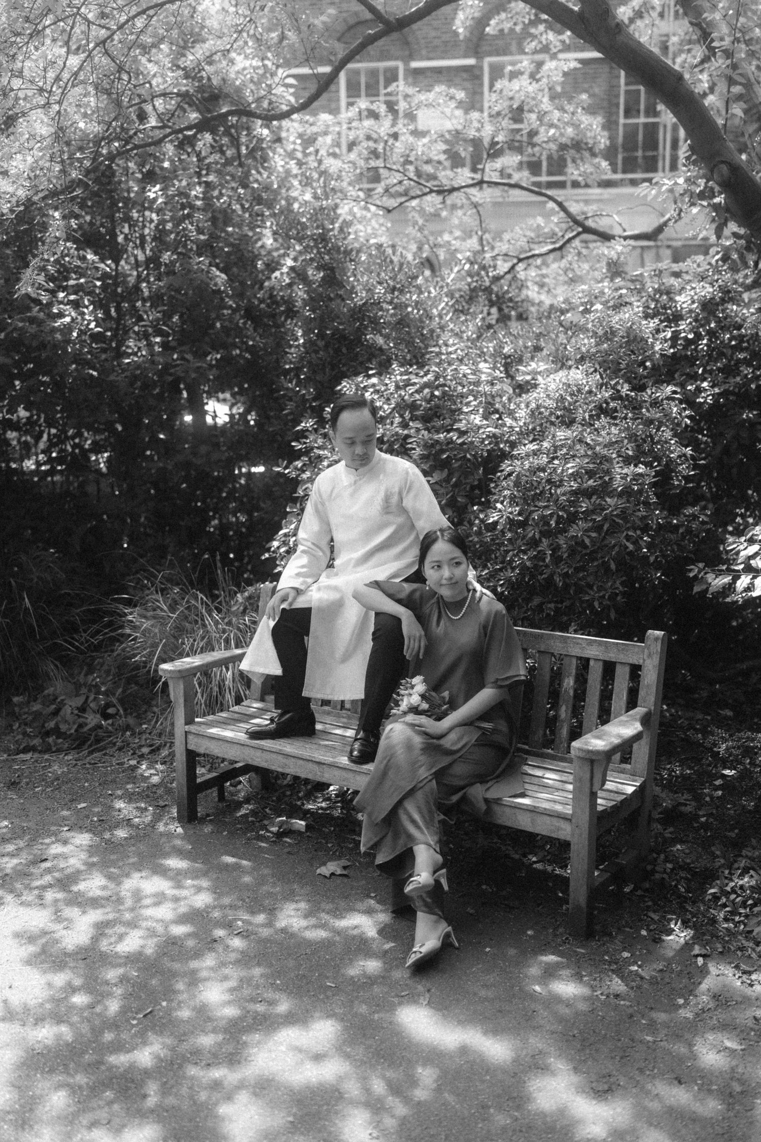 Two people pose on a wooden garden bench surrounded by lush greenery in a black and white photo.
