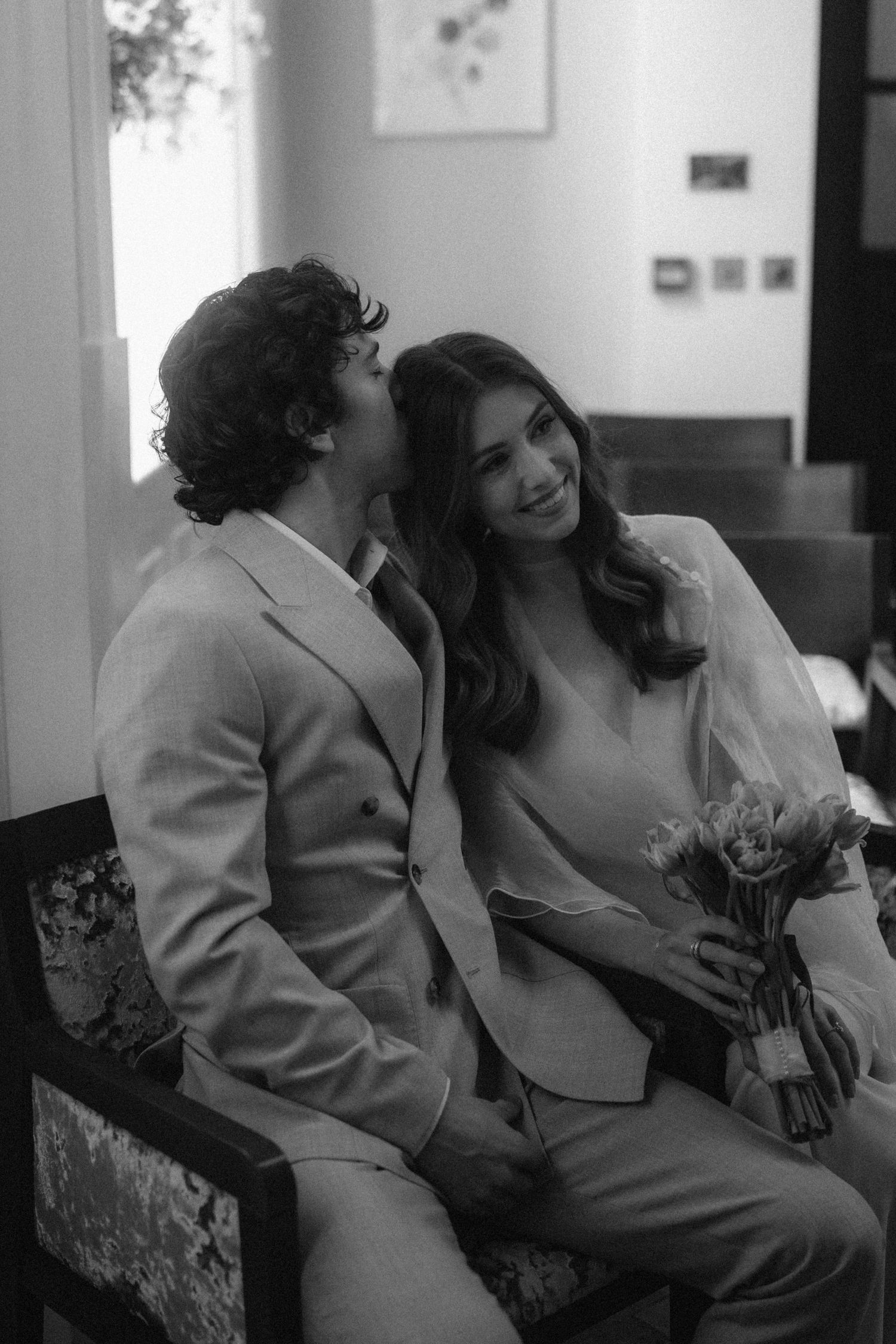 Black and white photo of a couple sitting together, man whispering to smiling woman holding a small bouquet.