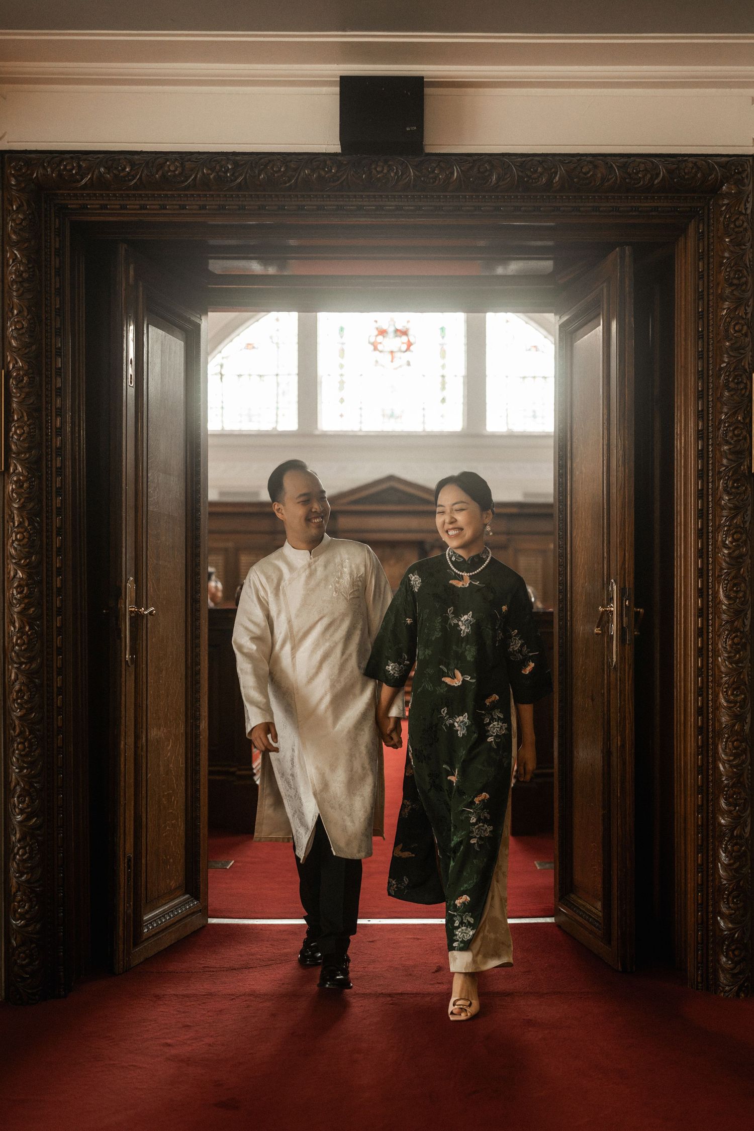 Couple in traditional Vietnamese ao dai attire walking through ornate wooden doors on red carpet.