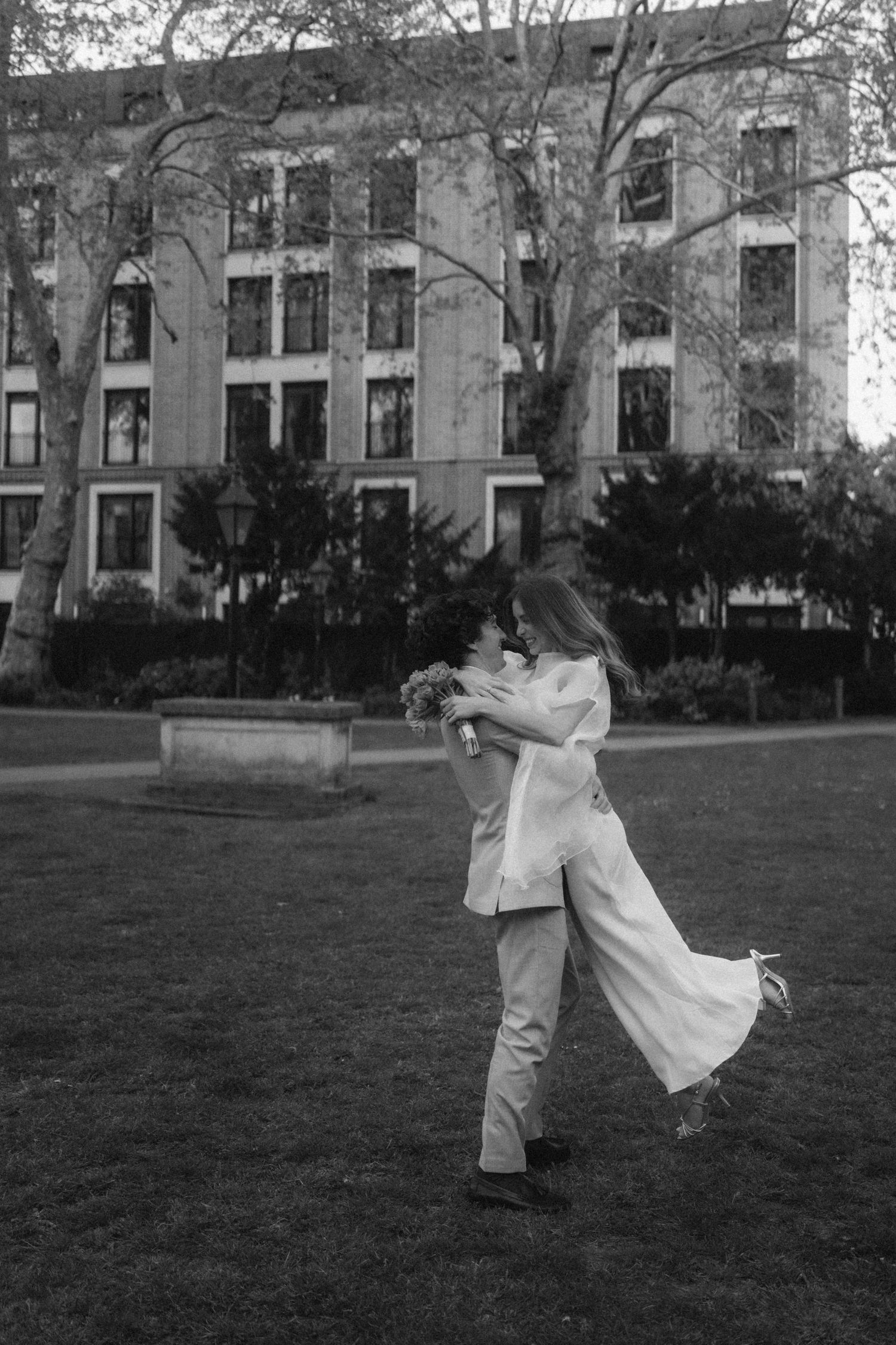Black and white photo of a groom lifting his bride in a park, both dressed in white wedding attire.