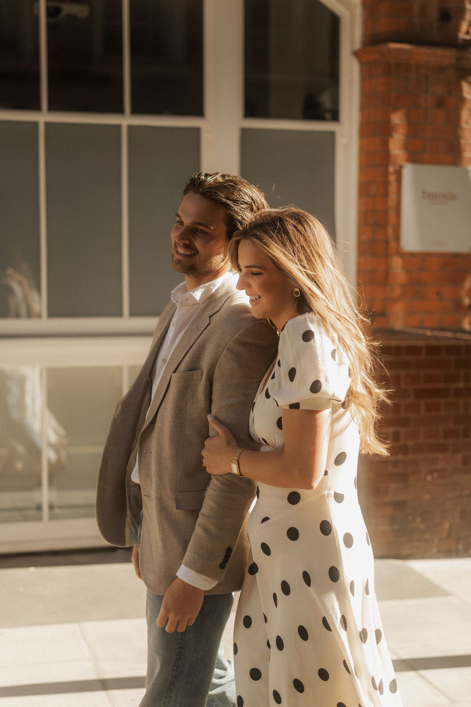 A smiling couple walks together outdoors, woman in polka dot dress embracing man in gray suit near brick building.